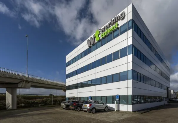 Een modern kantoorgebouw met de naam "Working Talent" en meerdere geparkeerde auto's ervoor, naast een viaduct.