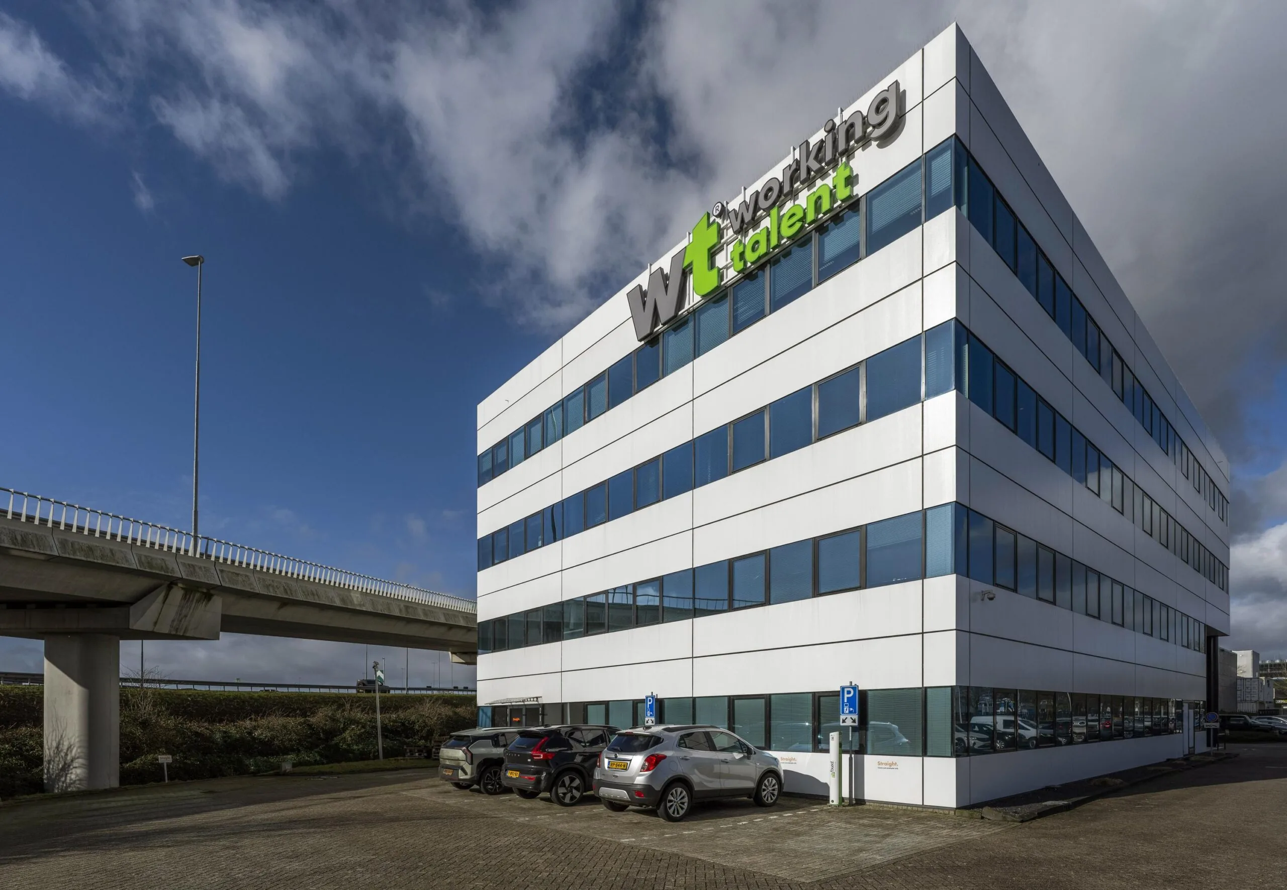 Een modern kantoorgebouw met de naam "Working Talent" en meerdere geparkeerde auto's ervoor, naast een viaduct.