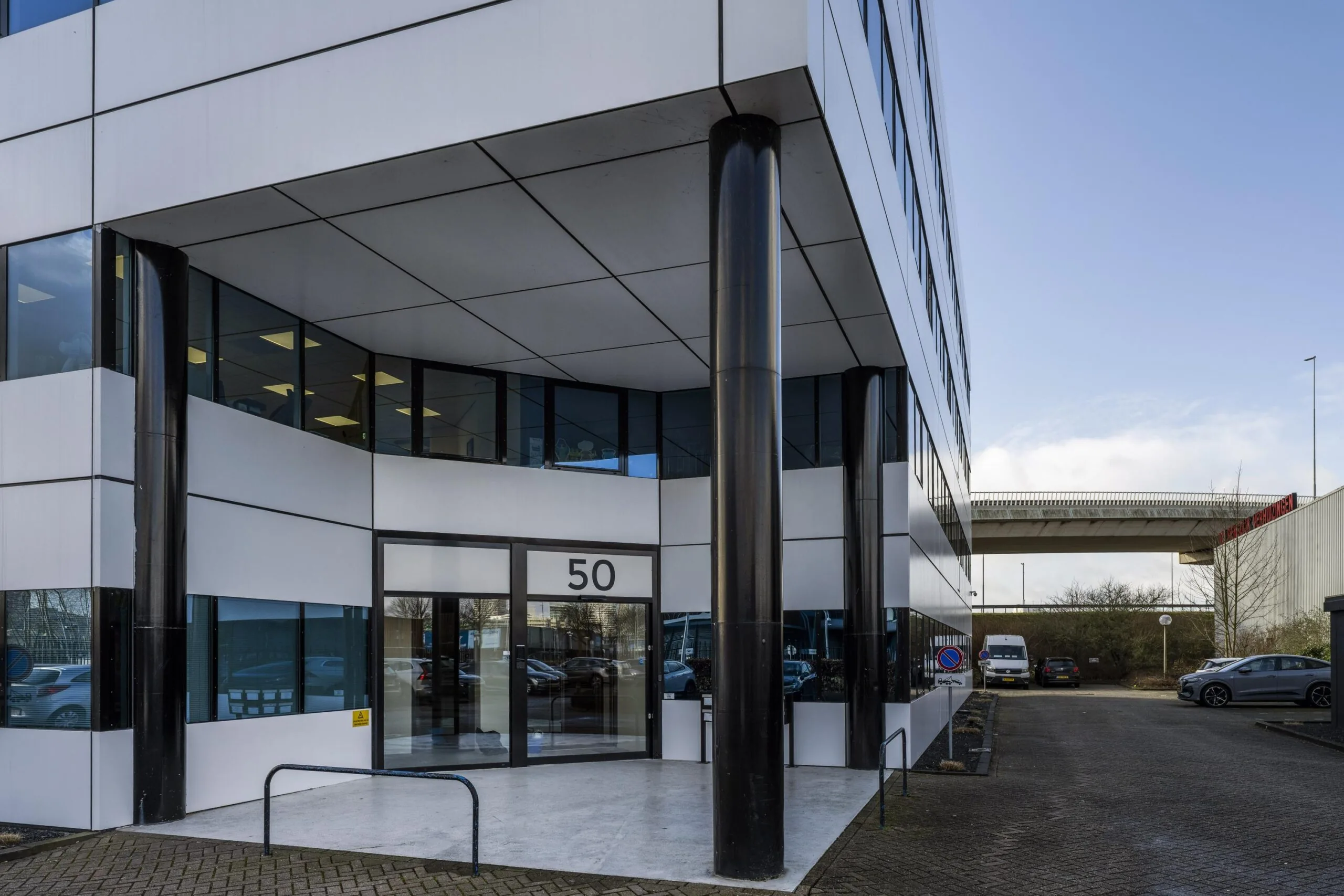 Een modern kantoorgebouw met het nummer 50 bij Kuiperbergweg, omgeven door parkeerplaatsen en een snelweg in de achtergrond.