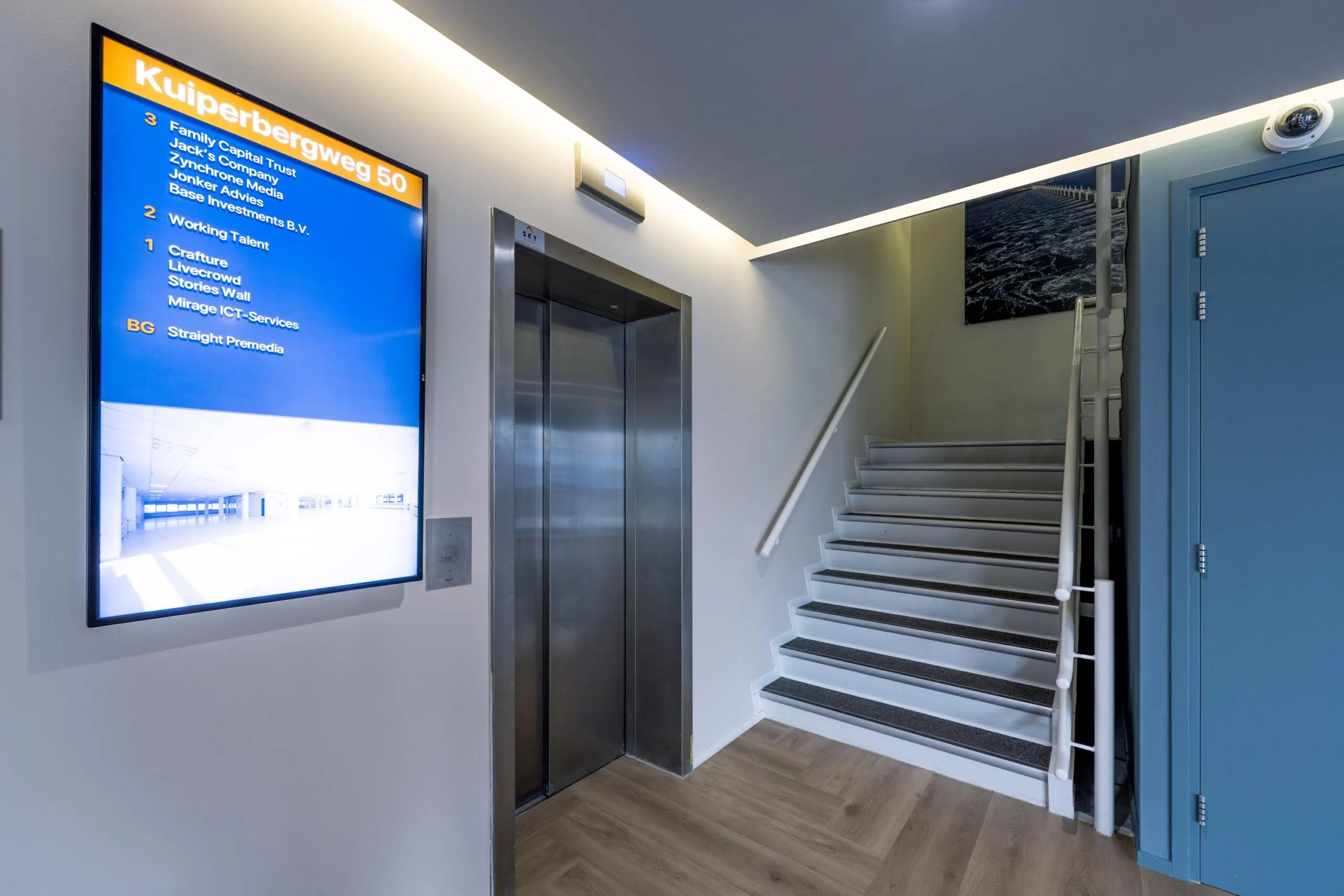 Een goed verlichte hal met een lift en een informatiebord dat de bedrijfsnamen op Kuiperbergweg 50 toont.