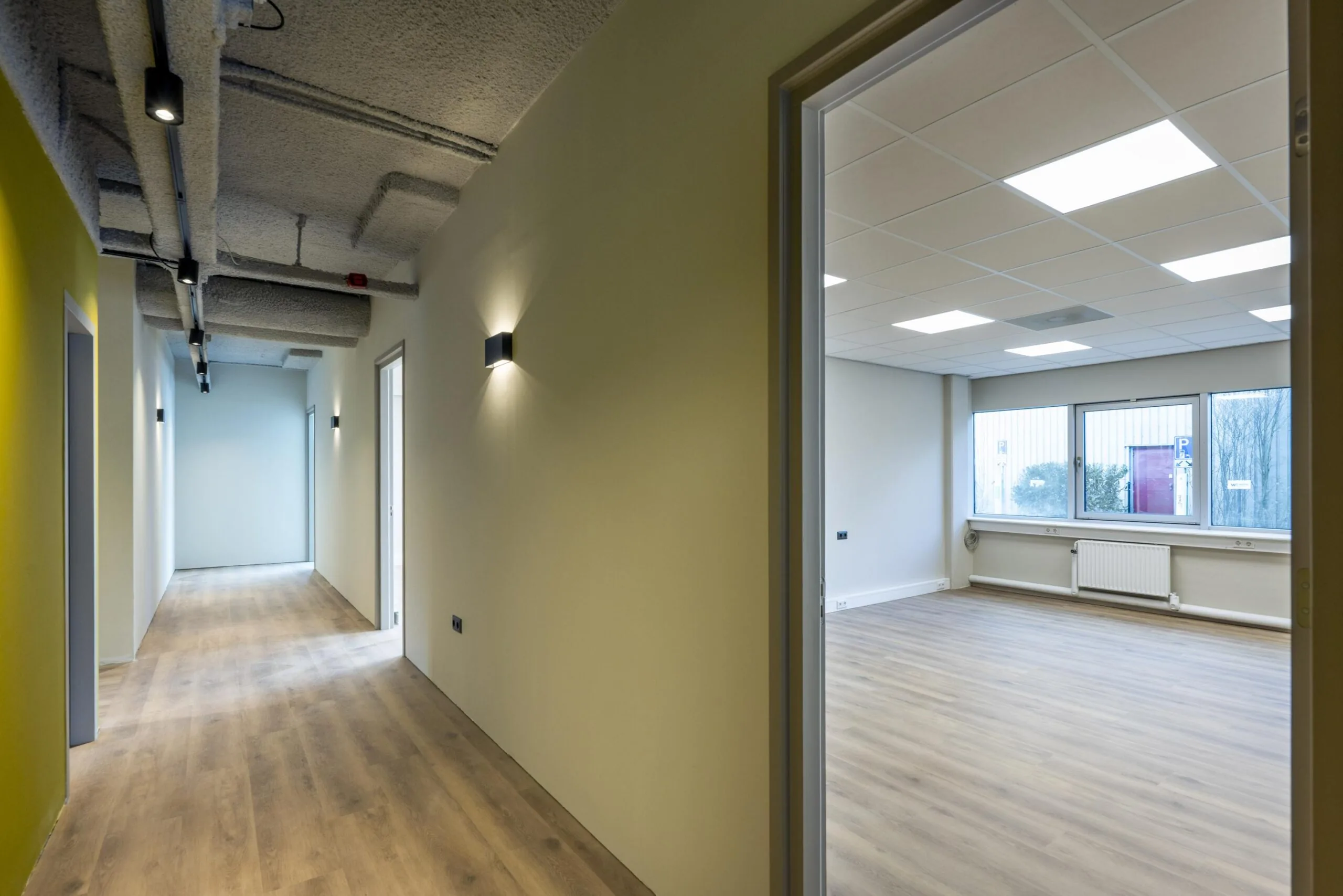 Een moderne, lege kantoorruimte met licht houten vloer en grote ramen gezien vanuit een gang.