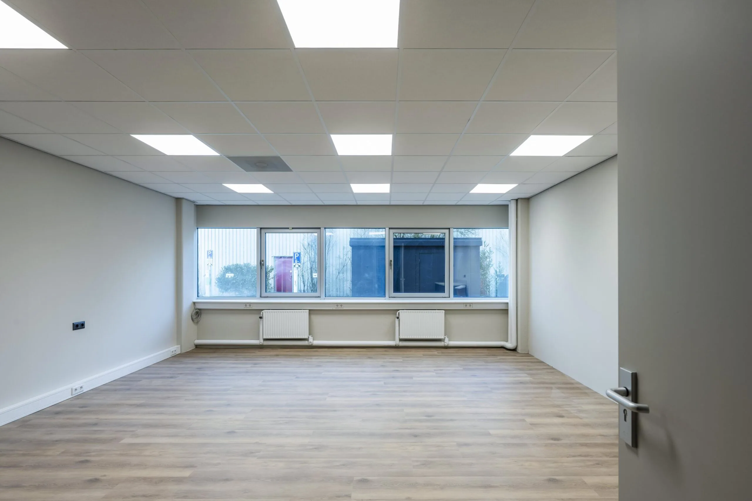 Lege kantoorruimte met houten vloer, grote ramen en een verlaagd plafond met inbouwverlichting.