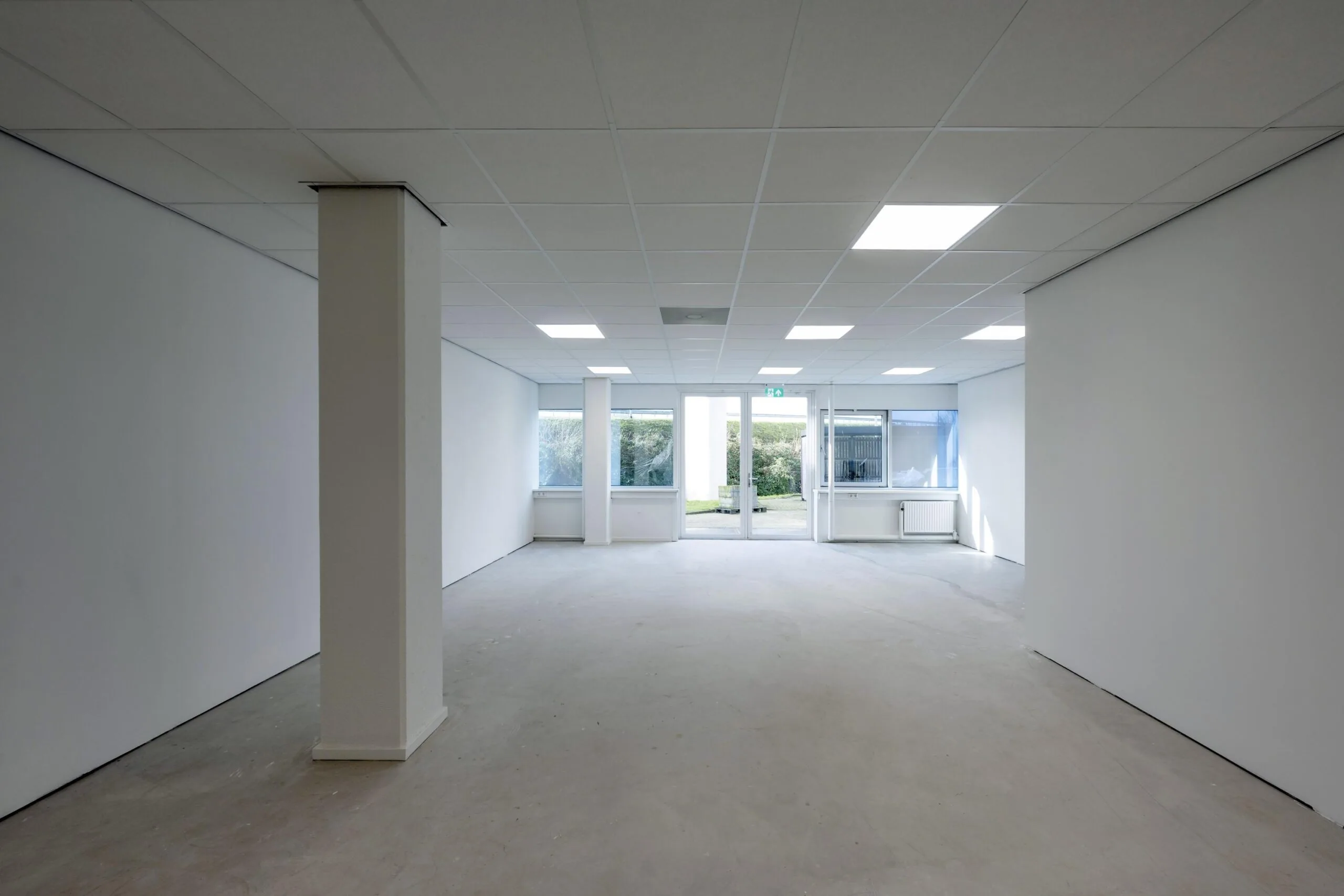 Een lege kantoorruimte met witte muren, verlaagd plafond en grote ramen aan het einde.