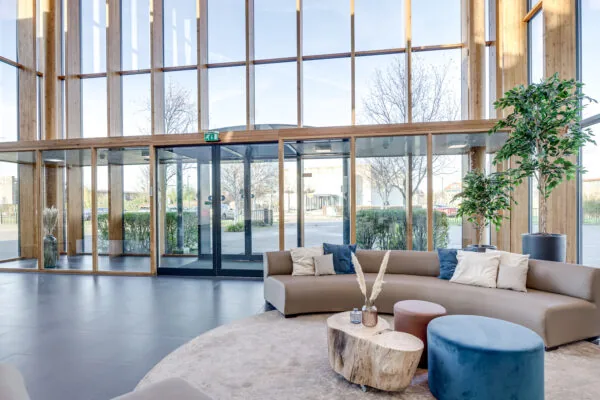 Een moderne ruimte met hoge ramen, een rond beige bankstel met kussens en een houten salontafel met decoratieve vazen.