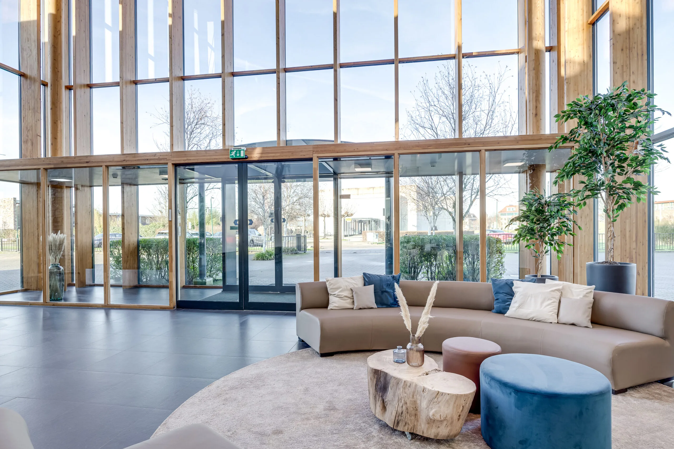 Een moderne ruimte met hoge ramen, een rond beige bankstel met kussens en een houten salontafel met decoratieve vazen.