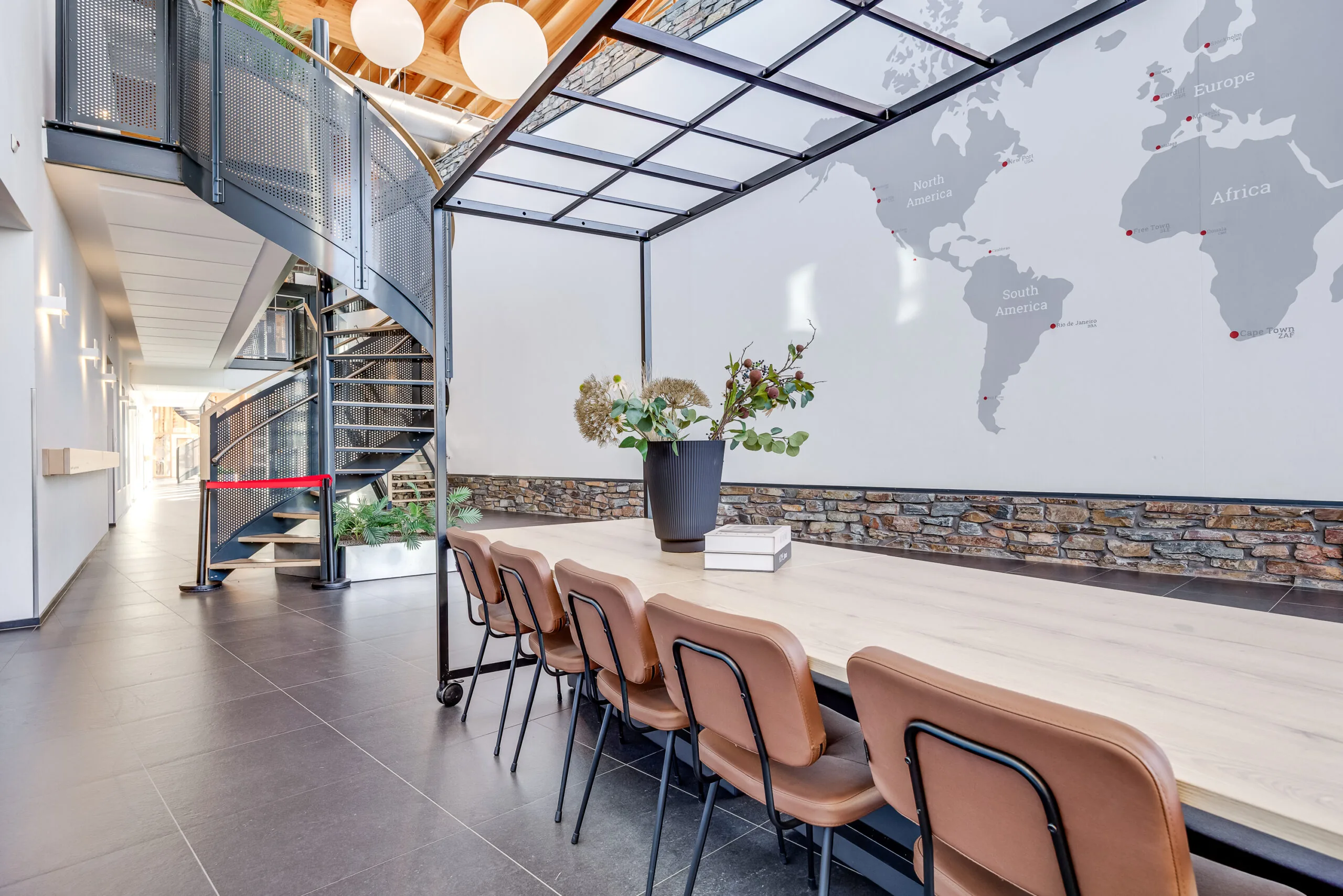 Een moderne ruimte met een houten tafel, leren stoelen, een wenteltrap en een wereldkaart op de muur.