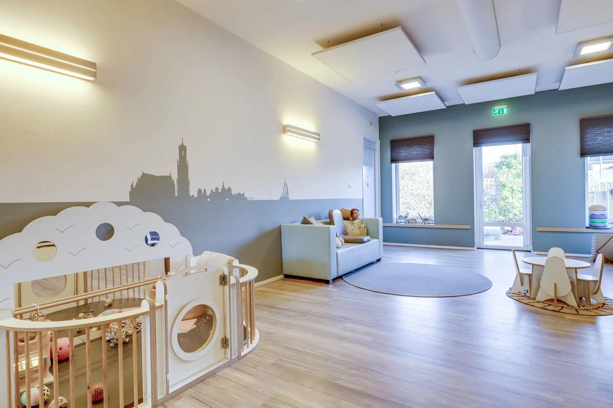 Een lichte en ruime speelkamer met kindermeubilair en een stadssilhouet op de muur.
