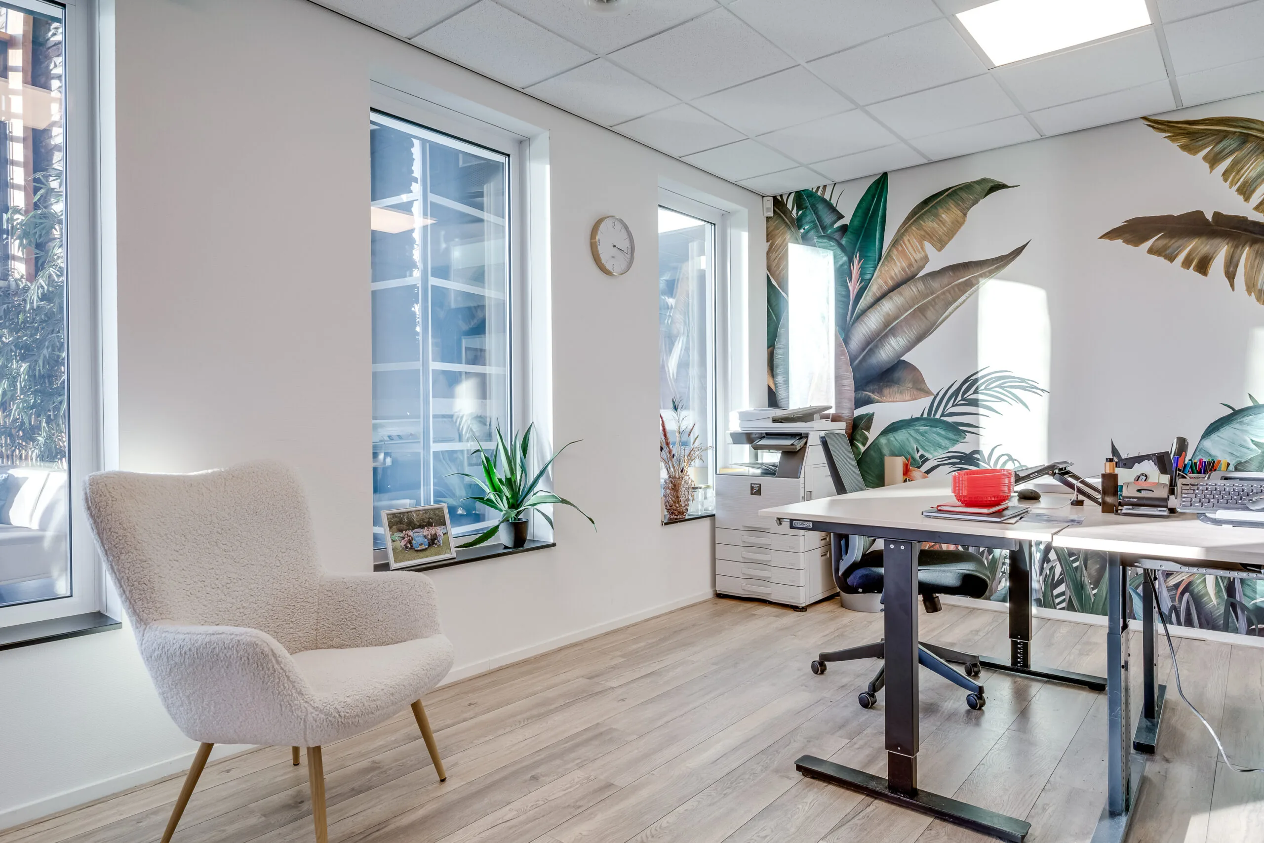 Een modern kantoor met een witte fauteuil, grote ramen, en tropisch behang.