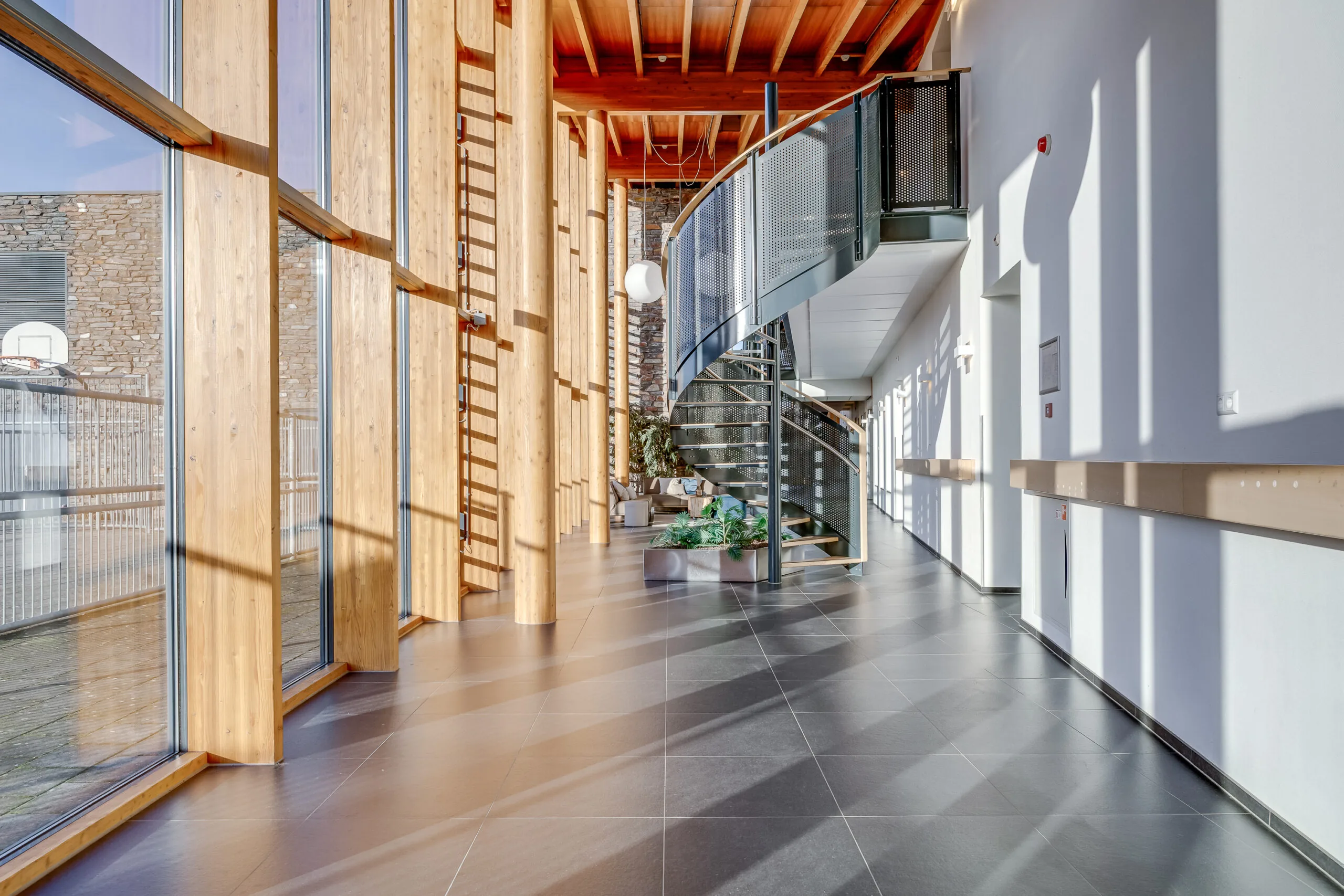 Een moderne kantoorhal met een glazen wand, houten steunbalken en een wenteltrap.