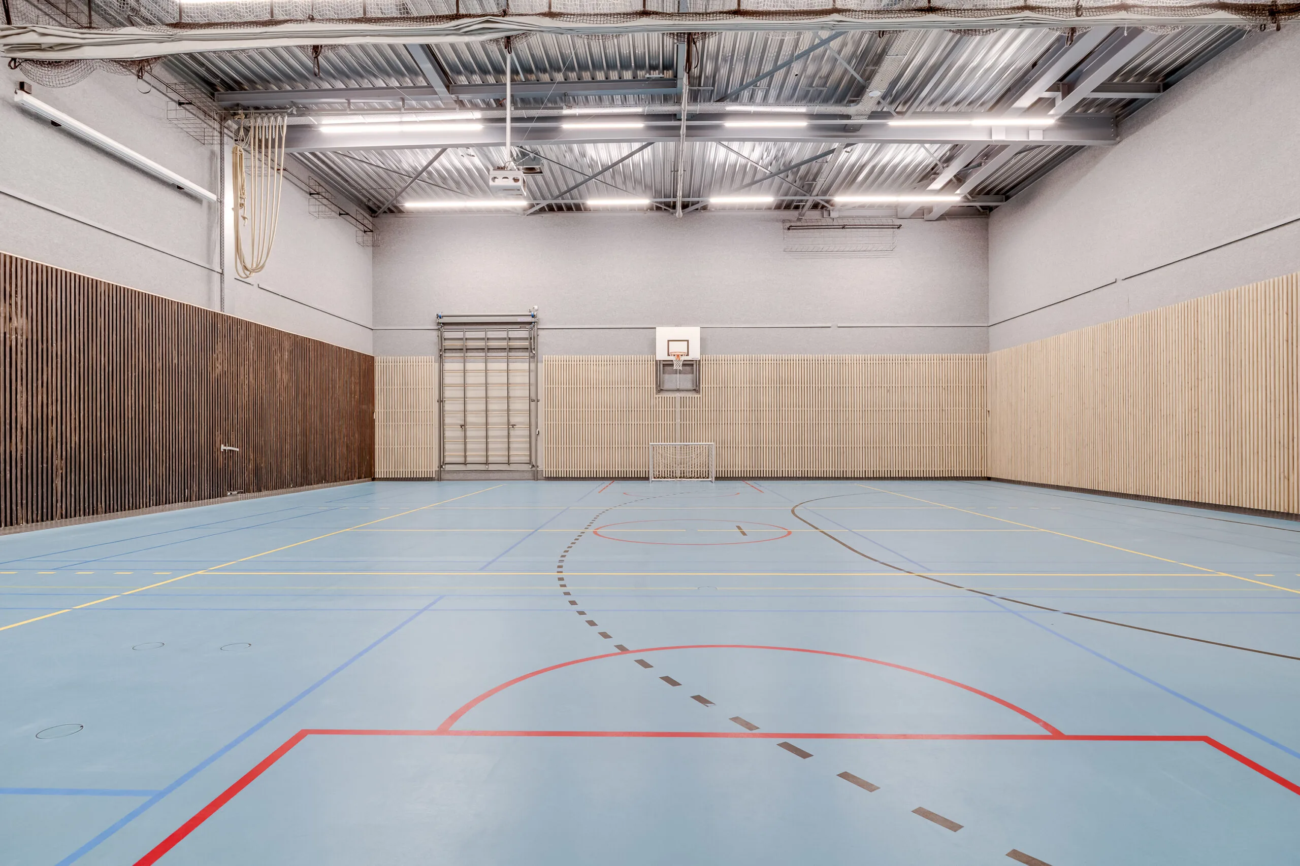 Een lege sportzaal met blauwe vloer en basketbalbord aan de muur.