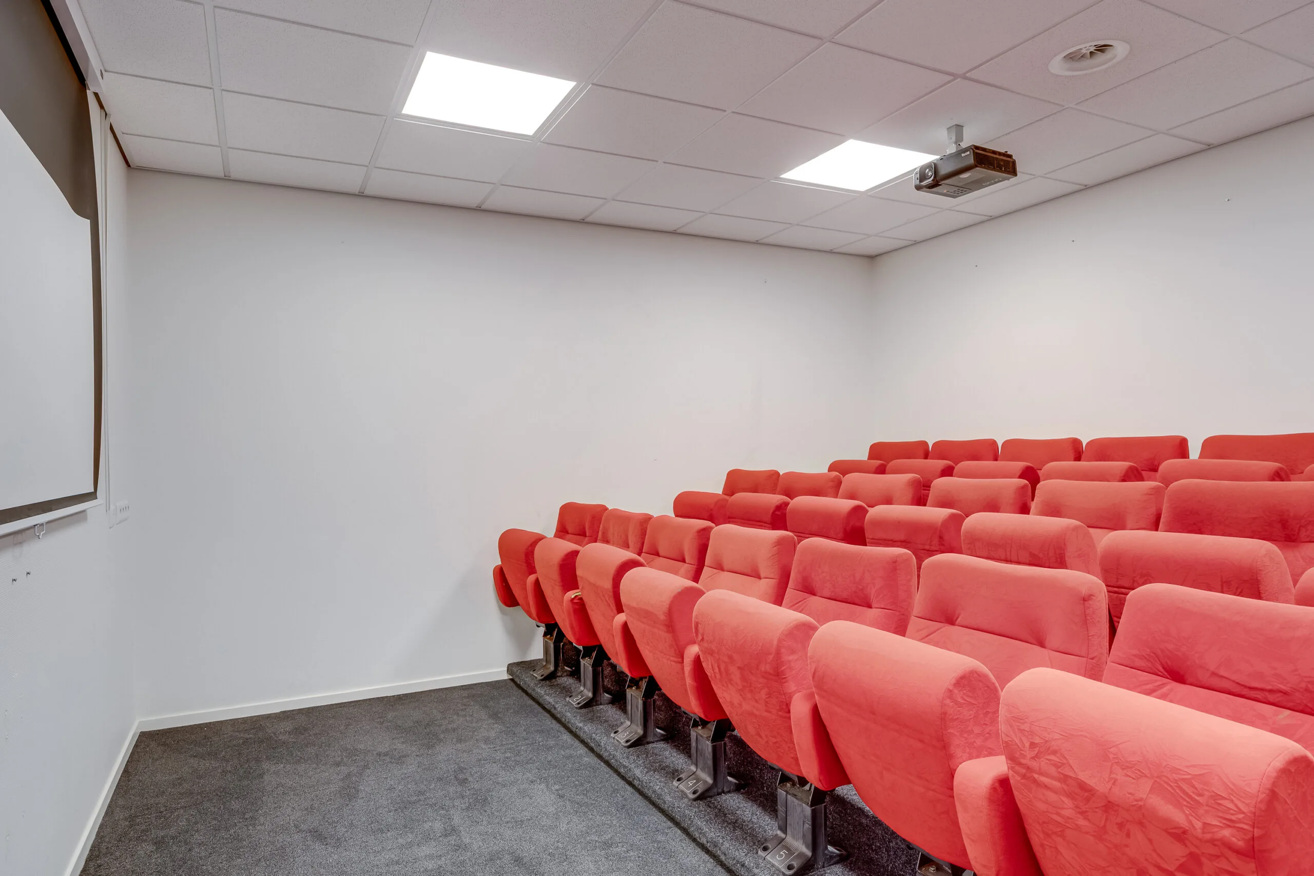 Een lege conferentiezaal met rode stoelen, gericht naar een projectorscherm.