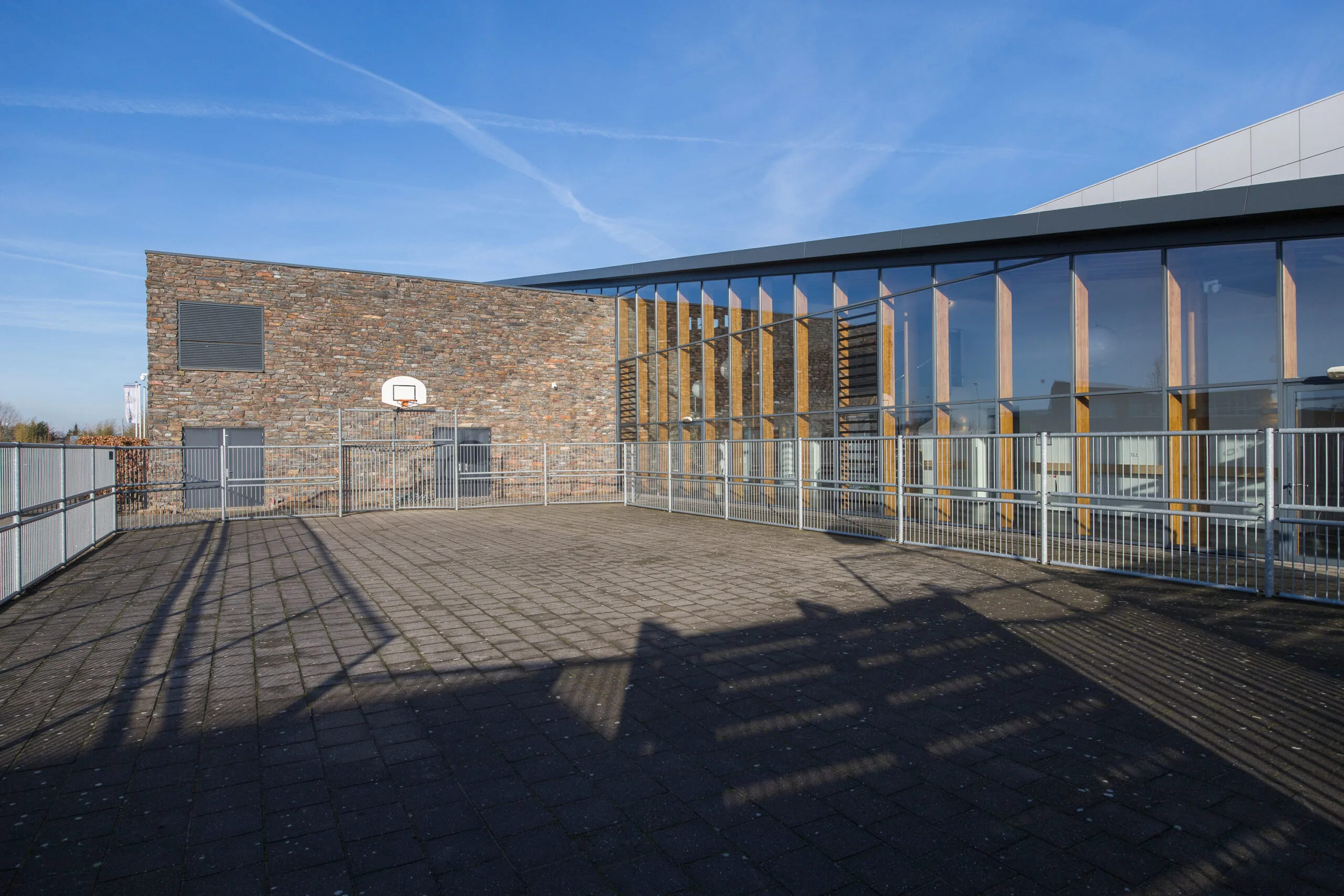 Een moderne speelplaats met een bakstenen muur en een glazen gevel, inclusief een basketbalring.