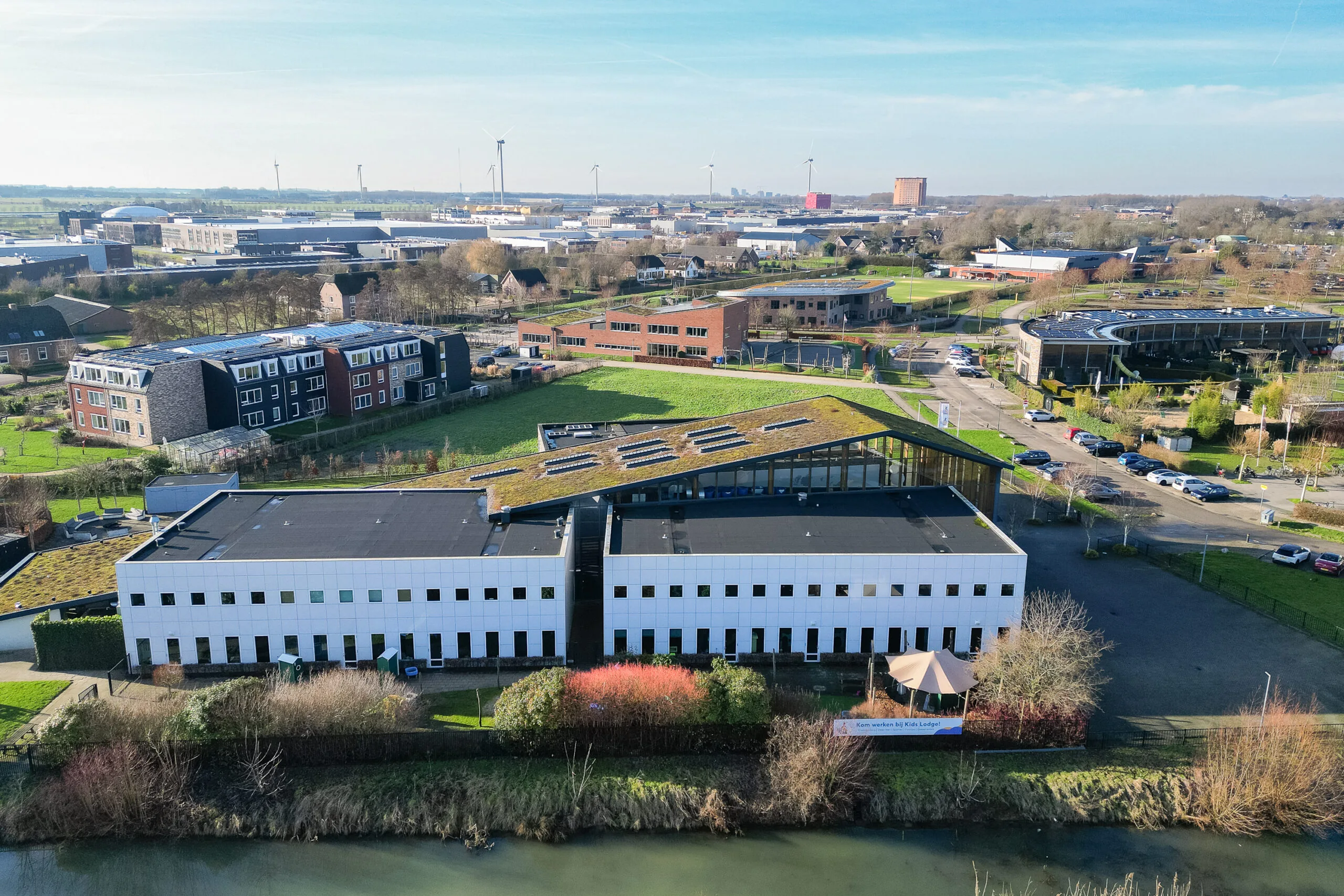 Luchtfoto van een gebouw met een groen dak en omliggende gebouwen en weilanden in een landelijke omgeving.