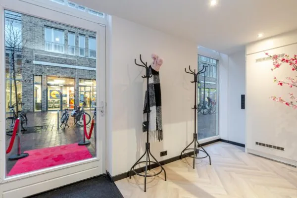 Een interieur met kapstokken, een roze tapijt bij de deur en uitzicht op een winkelstraat.