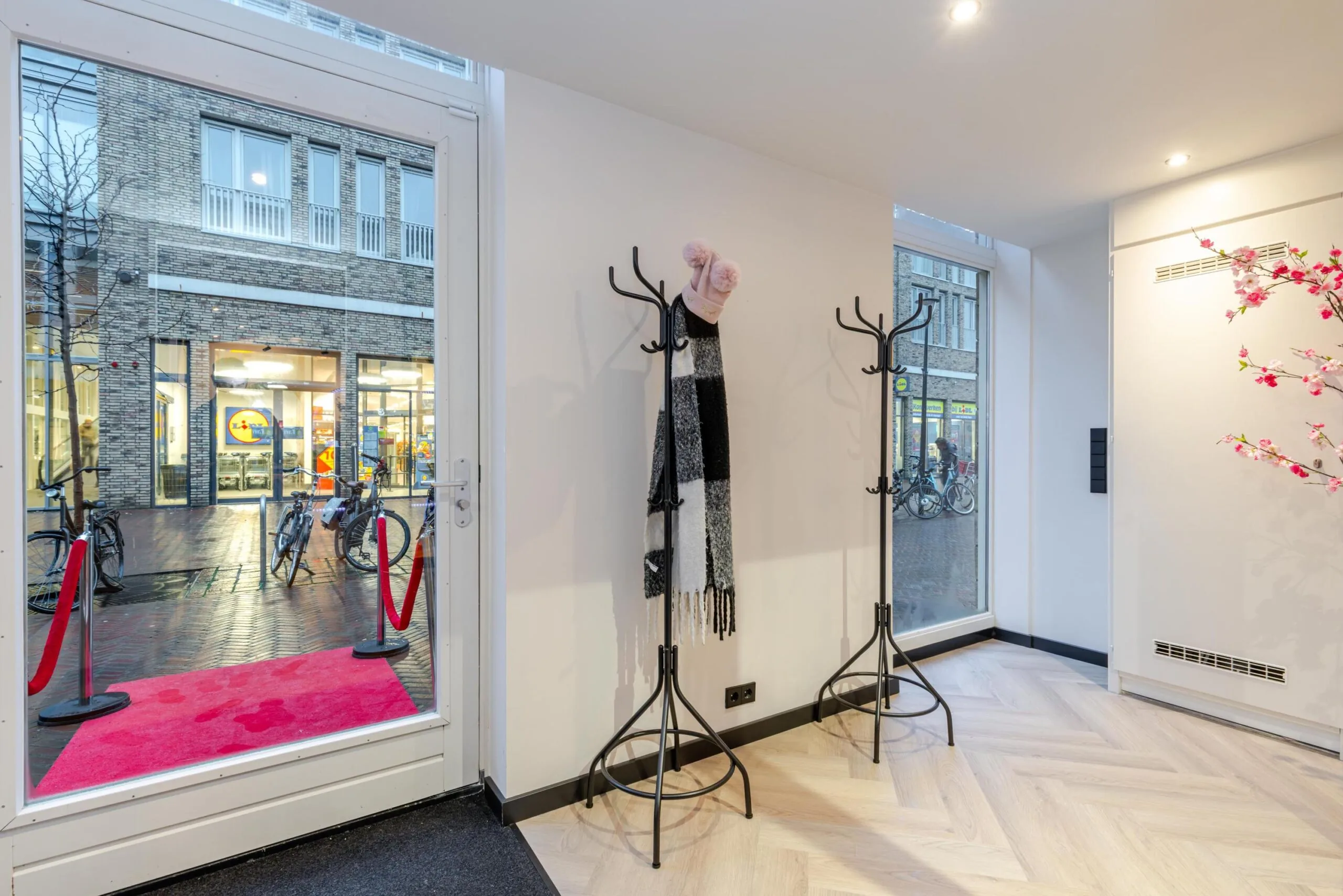 Een interieur met kapstokken, een roze tapijt bij de deur en uitzicht op een winkelstraat.