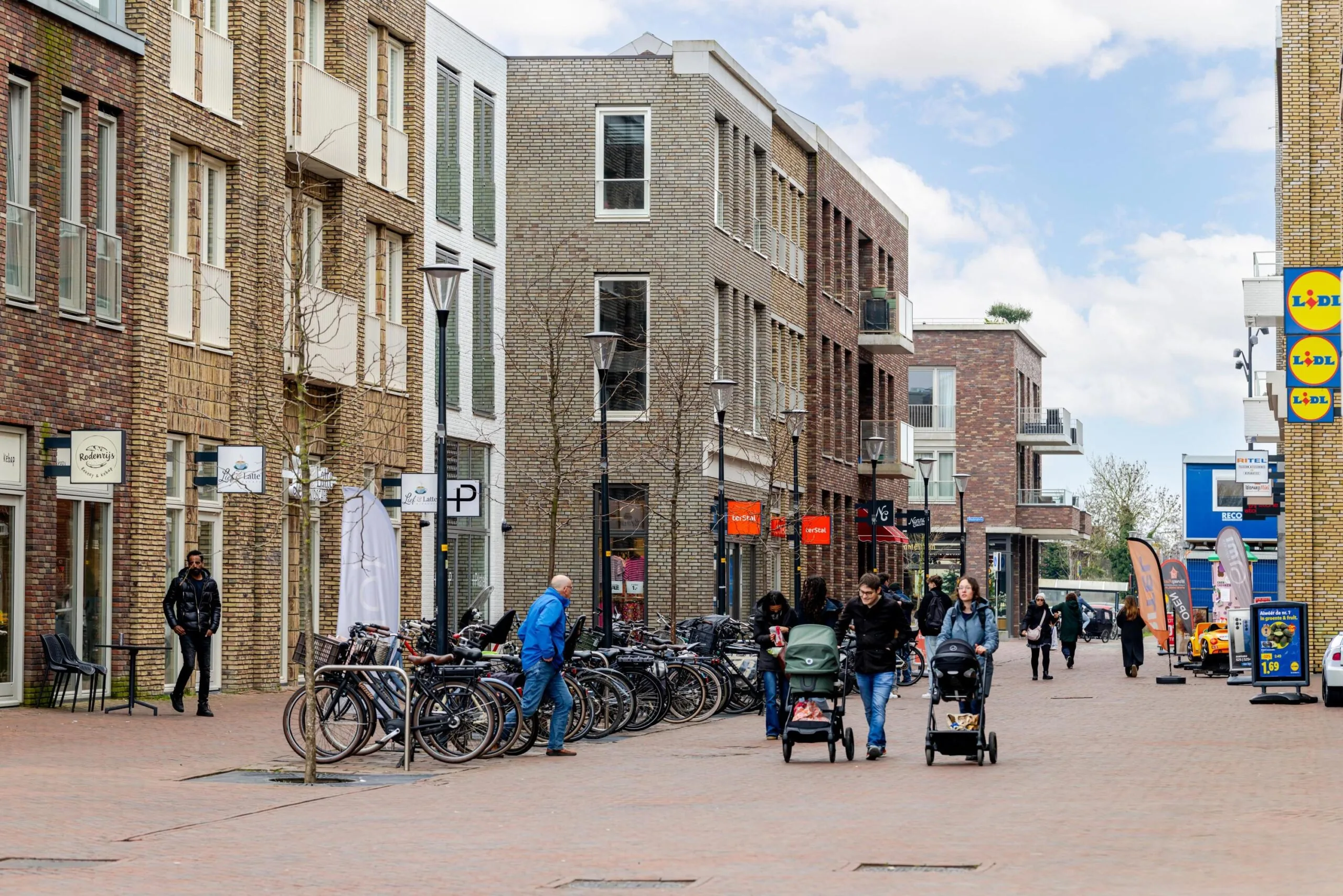 Een stadsstraat met bakstenen gebouwen, winkels en mensen die langs fietsen en kinderwagens lopen.