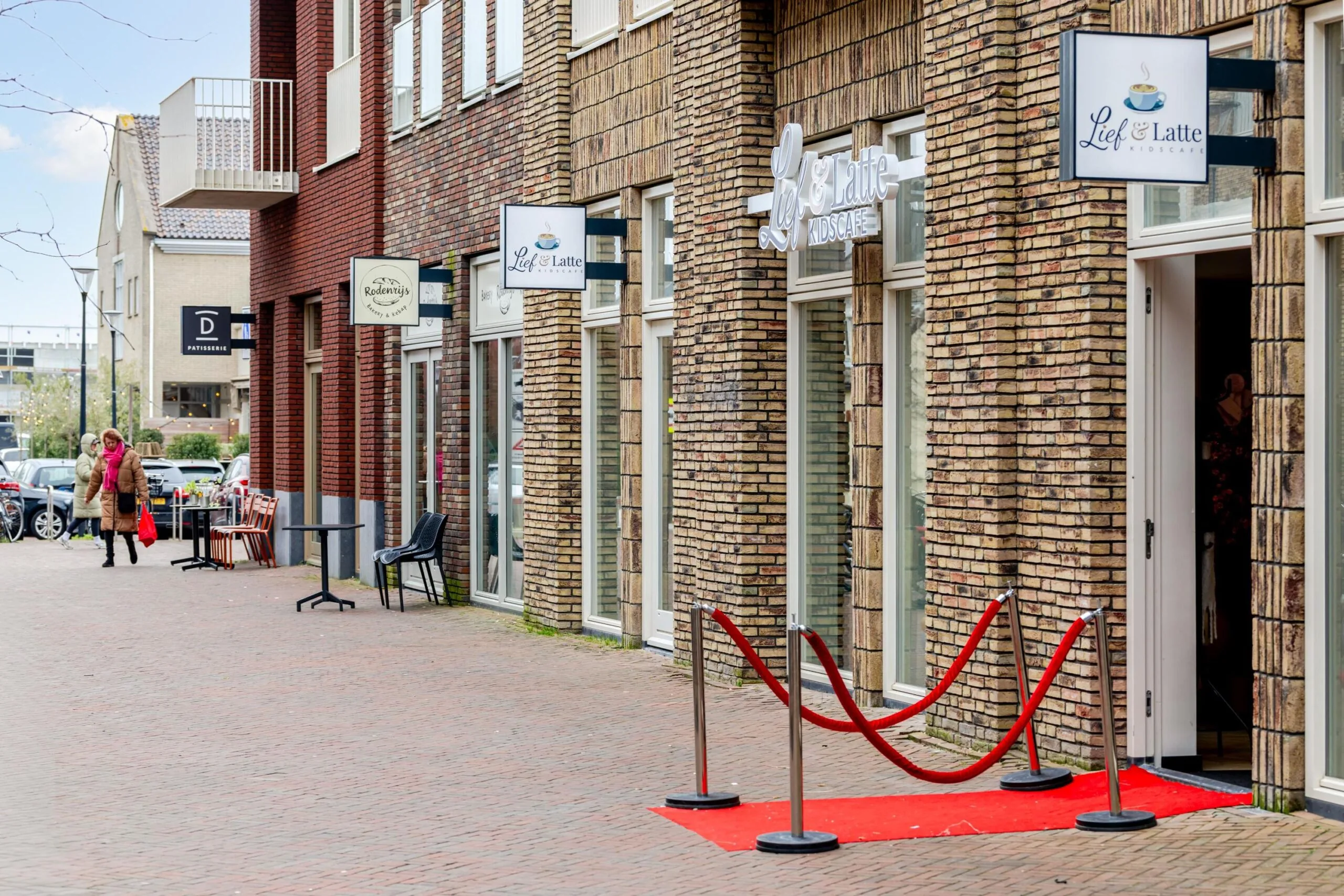 Een straatzicht van een rij winkels, waaronder "Lief & Latte Kidscafe", met een rode loper bij de ingang.
