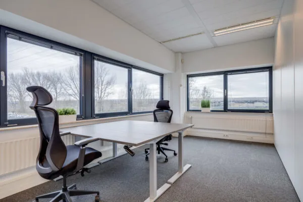 Een moderne, lichte kantoorruimte met twee zwarte bureaustoelen en grote ramen met uitzicht naar buiten.