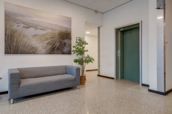 Een wachtruimte met een grijze bank, een groot natuurcanvas en een liftdeur aan de rechterkant.