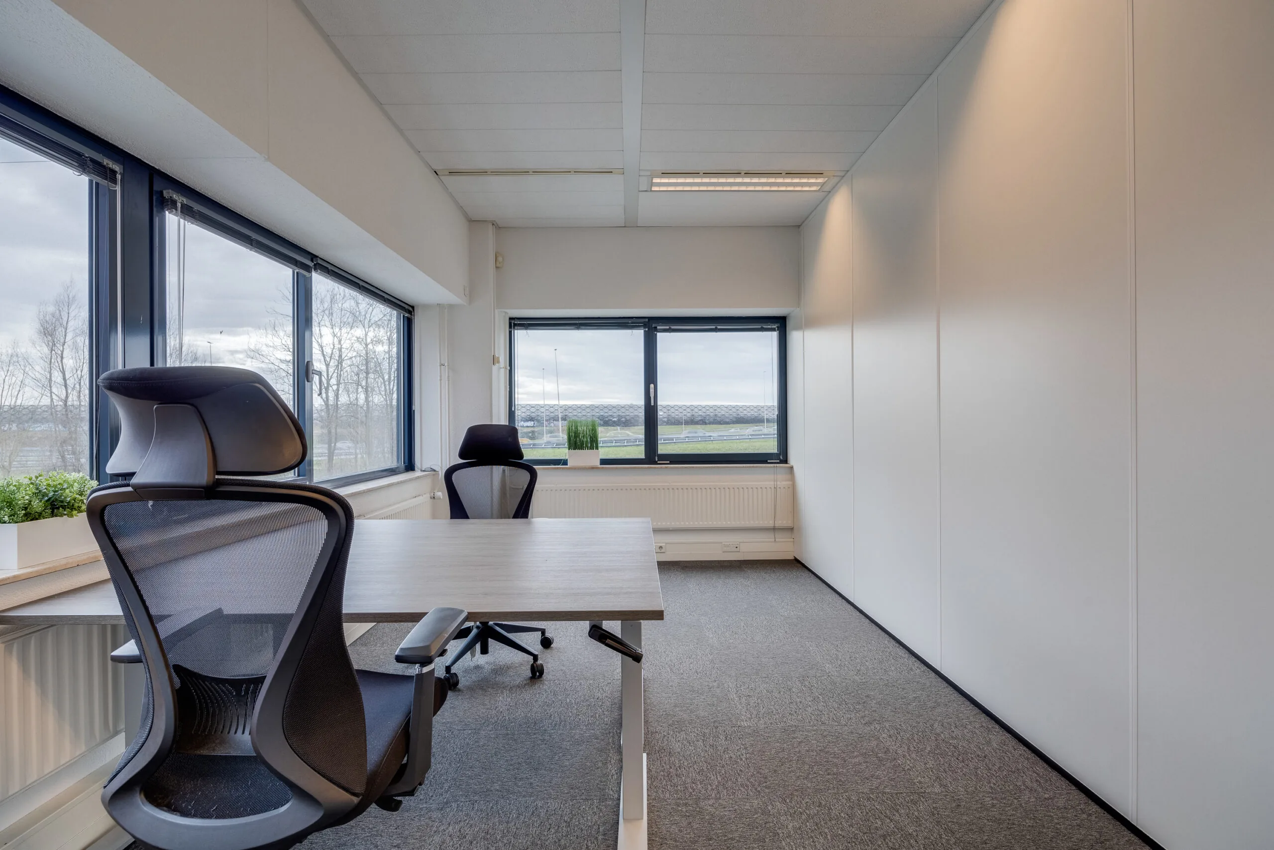 Een licht en modern kantoor met bureaus, stoelen en grote ramen met uitzicht naar buiten.