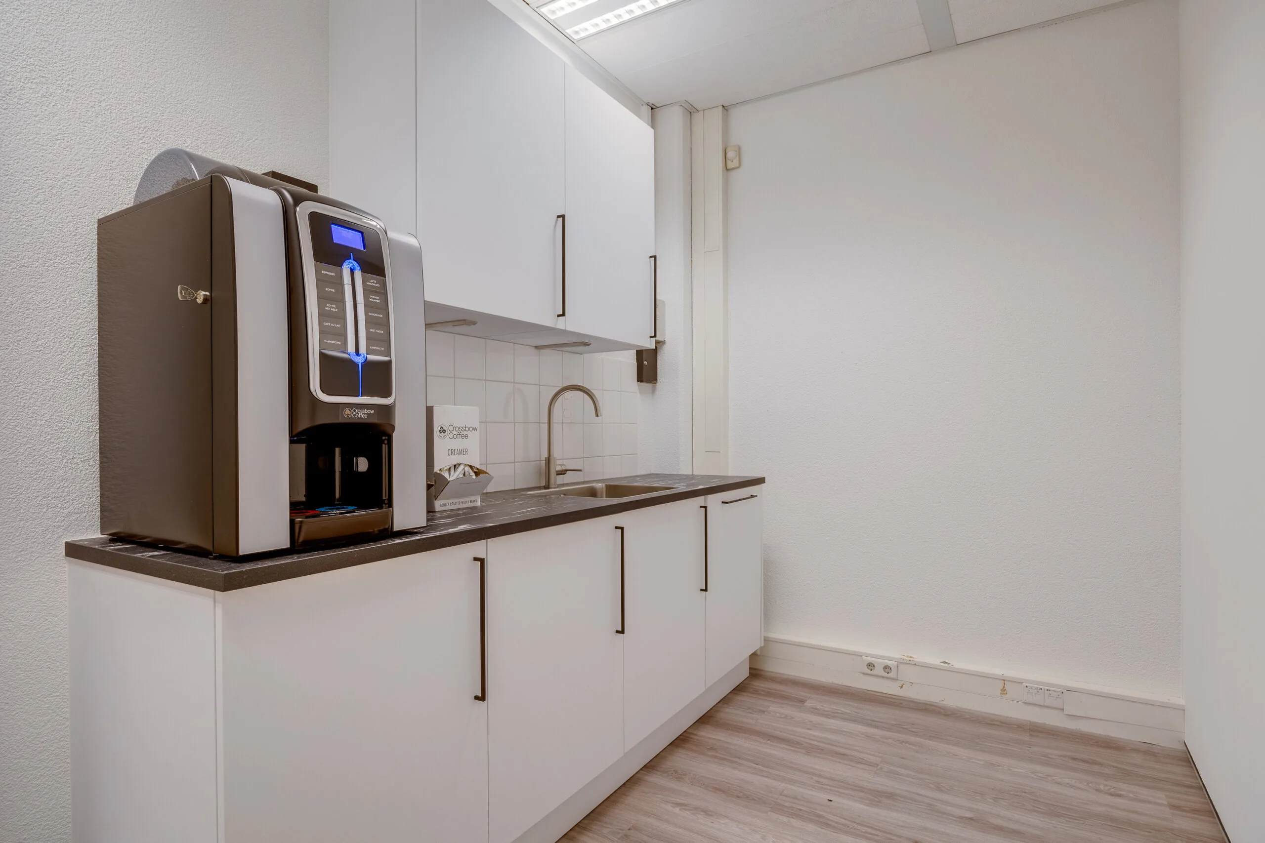 Een klein keukenblok met een koffiemachine en een gootsteen in een verder lege keukenruimte.