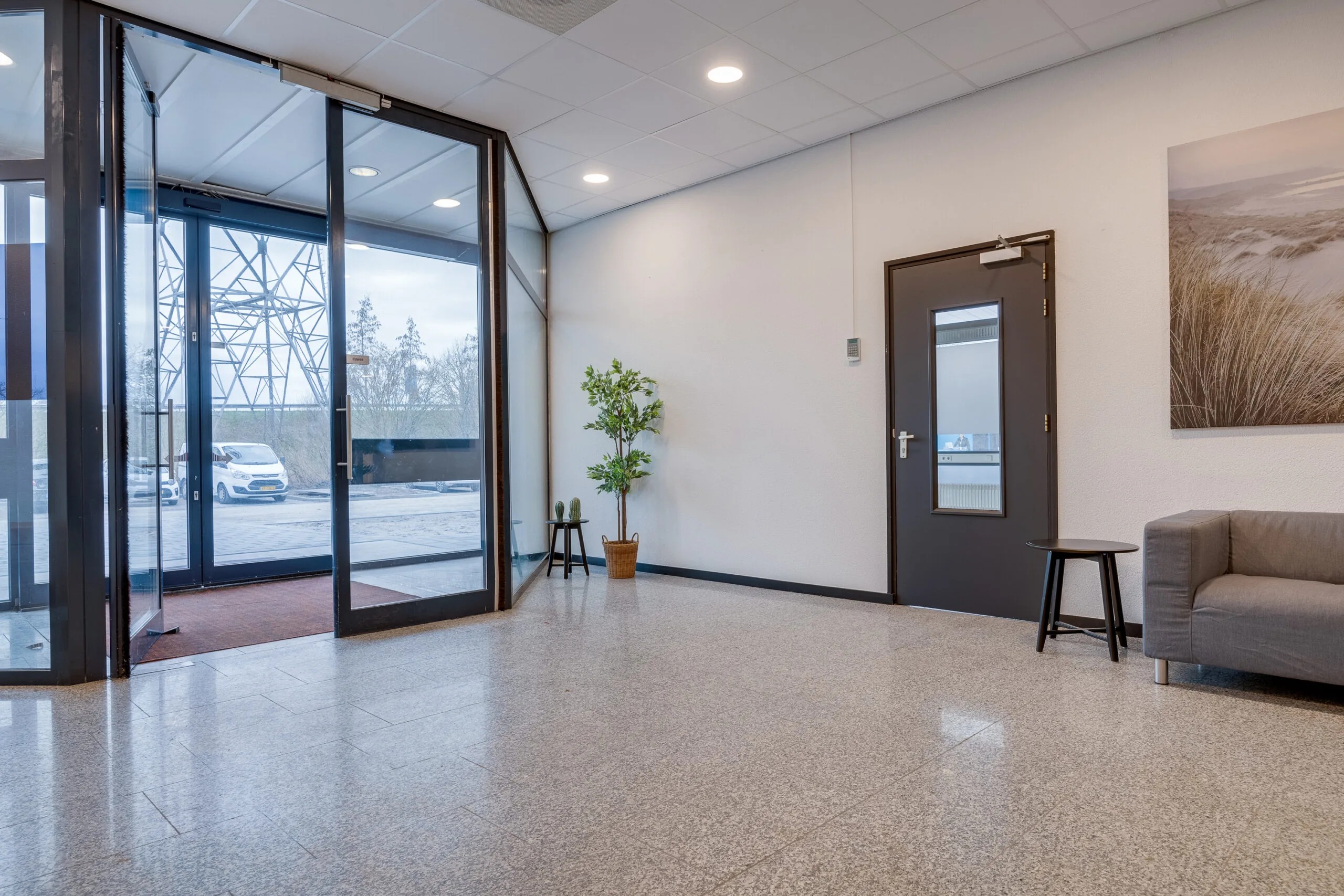 Een moderne, lichte ontvangstruimte met glazen deuren, een klein tafeltje, en een grijze bank.