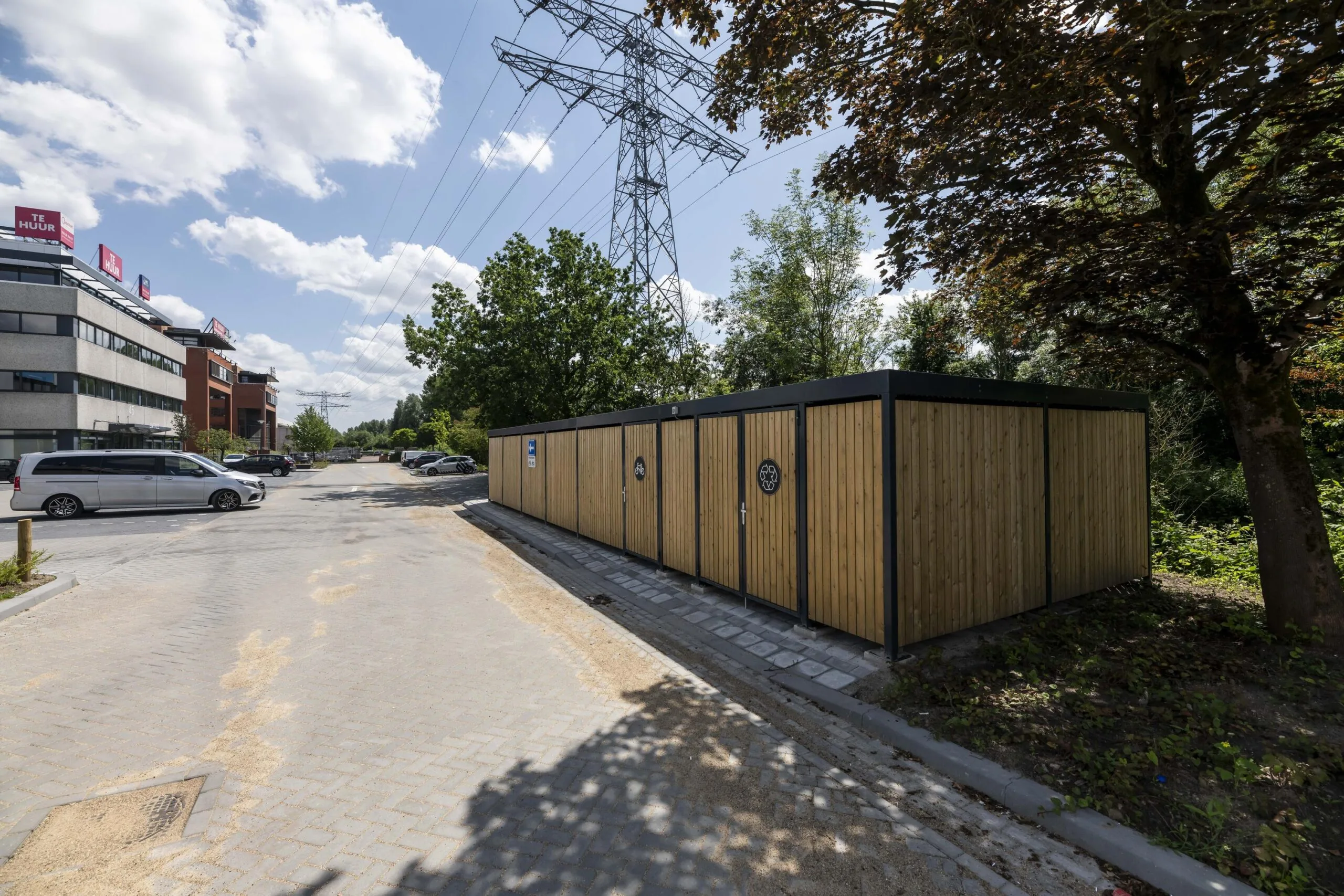 Een parkeerplaats naast een houten fietsenstalling en een hoogspanningsmast in een stedelijke omgeving.