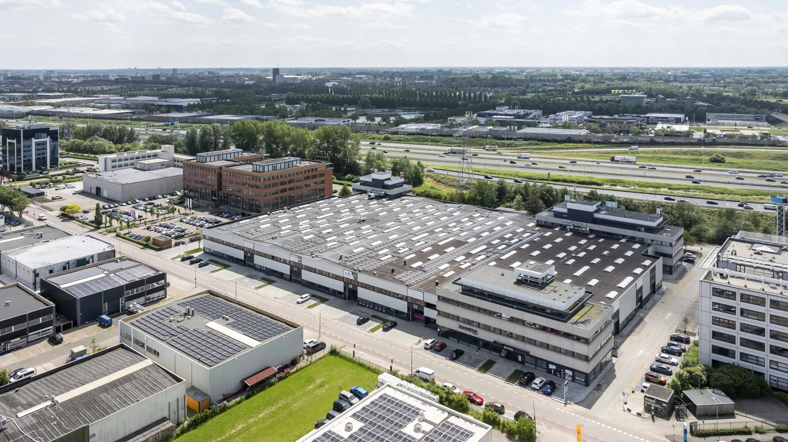 Luchtfoto van een bedrijventerrein met grote gebouwen en zonnepanelen op de daken, gelegen naast een snelweg.