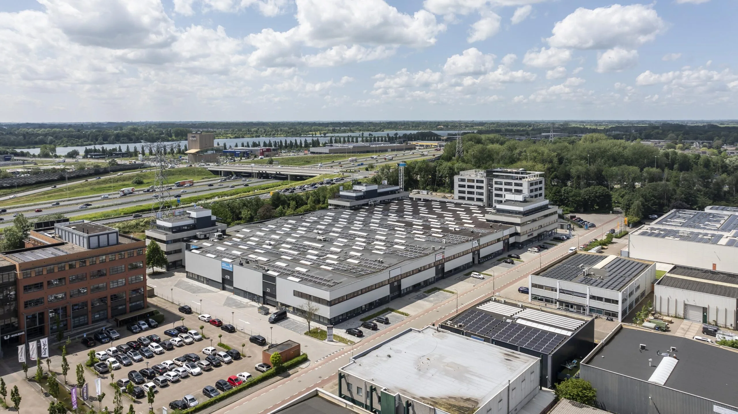 Luchtfoto van een industrieterrein met grote, platte daken en zonnepanelen, naast snelwegen en groene landschappen.