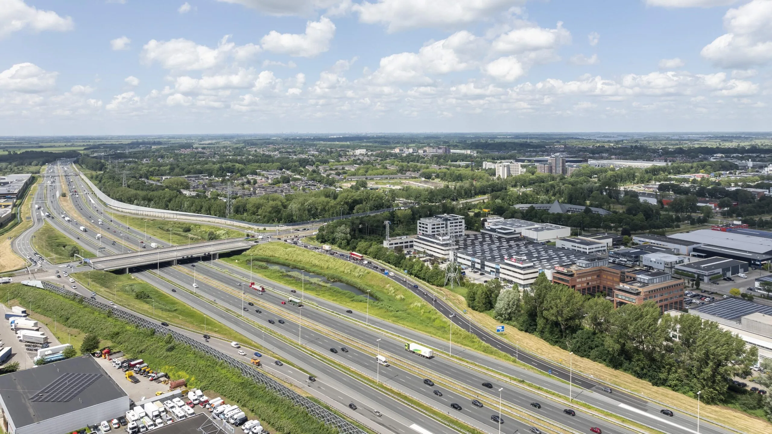 Luchtfoto van een snelweg en omliggende bebouwing en natuur, onder een licht bewolkte hemel.