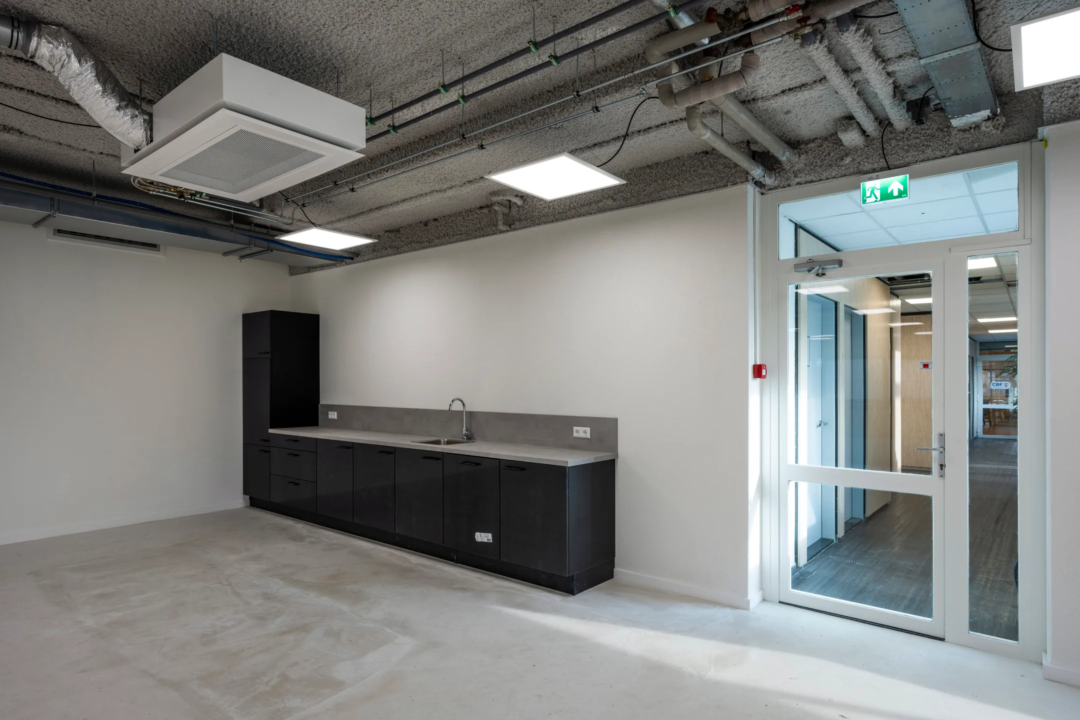 Een lege kantoorruimte met een kitchenette en een glazen deur naar een gang.