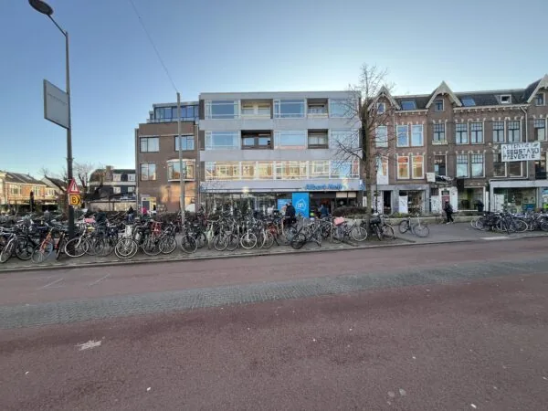 Straatbeeld van de Burgemeester Reigerstraat met geparkeerde fietsen en de gevels van een Albert Heijn supermarkt en omliggende gebouwen.