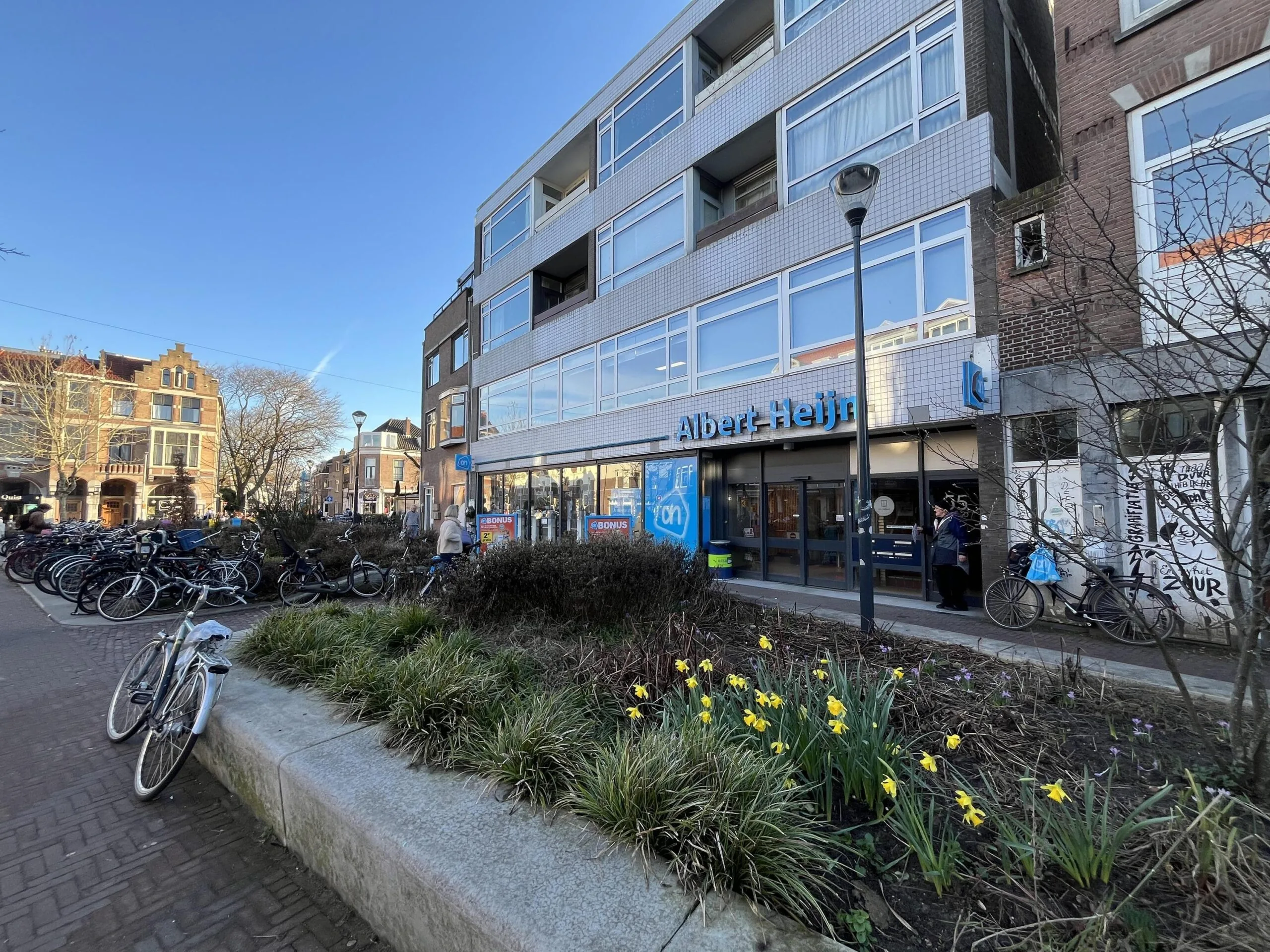 Een Albert Heijn supermarkt aan de Burgemeester Reigerstraat met fietsenrekken en bloeiende narcissen op de voorgrond.