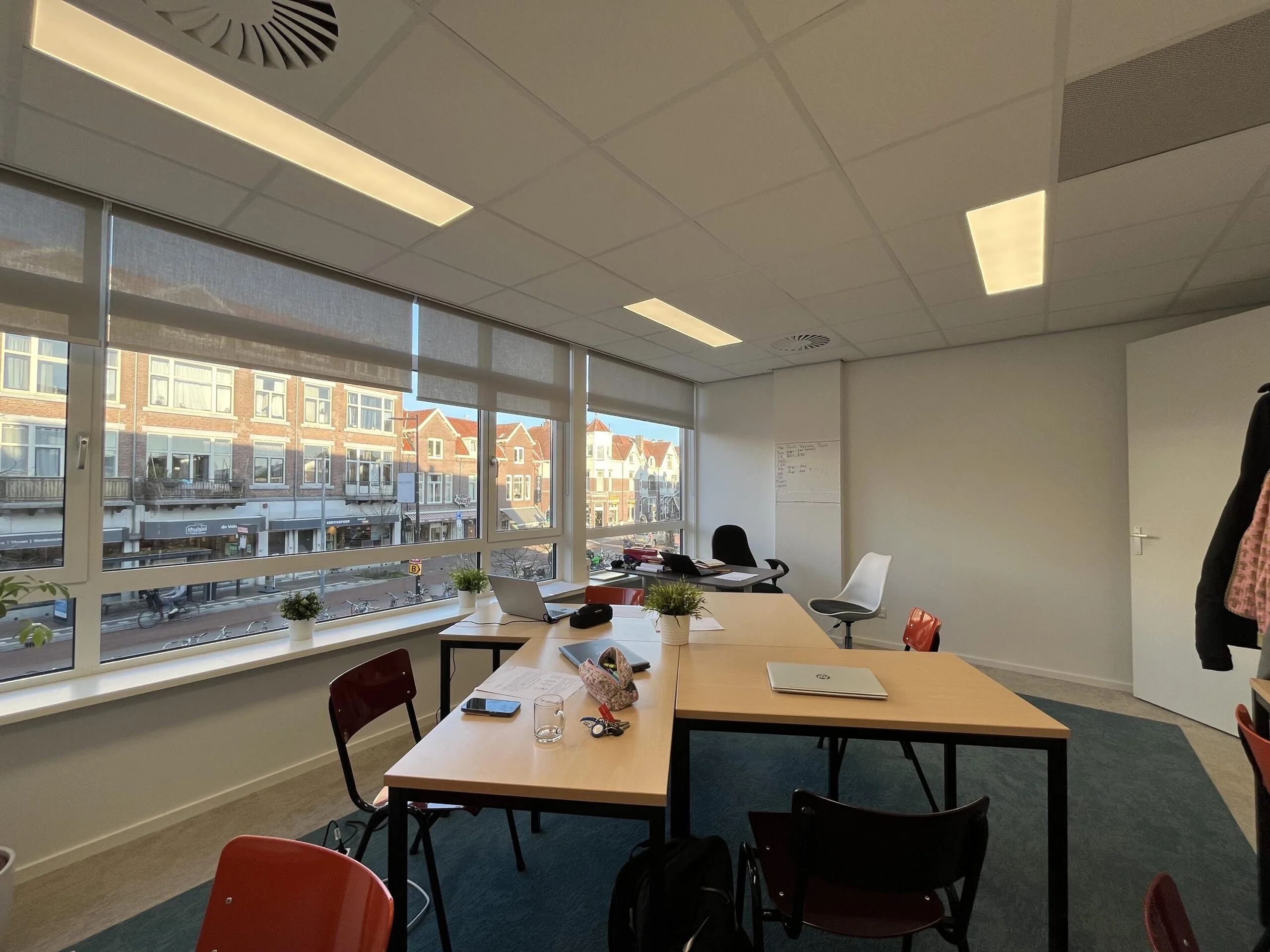Een moderne kantoorruimte met grote ramen, uitzicht op de Burgemeester Reigerstraat, en meerdere bureau's met stoelen.
