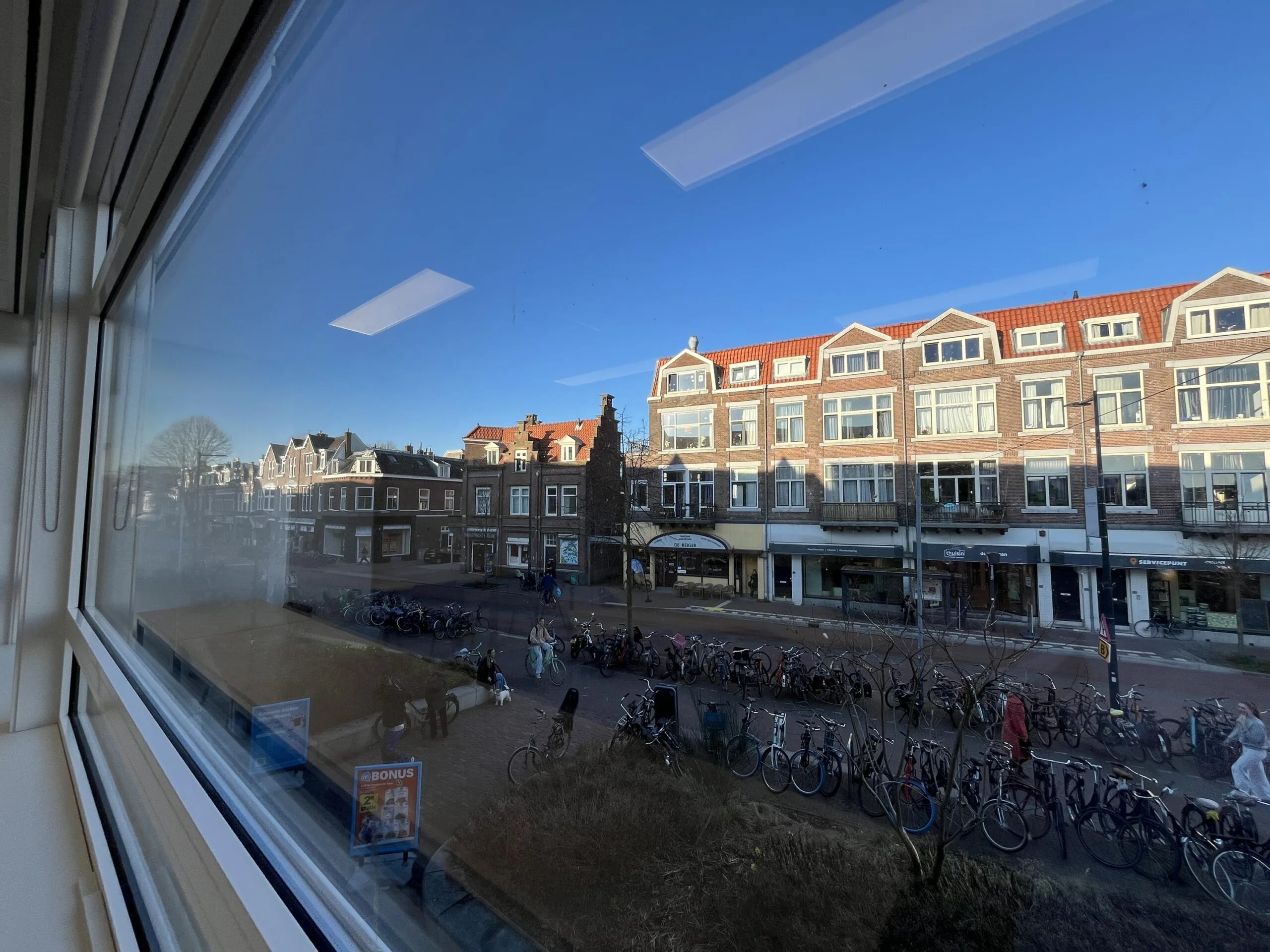 Uitzicht vanuit een raam op de Burgemeester Reigerstraat met geparkeerde fietsen en historische gebouwen op een zonnige dag.