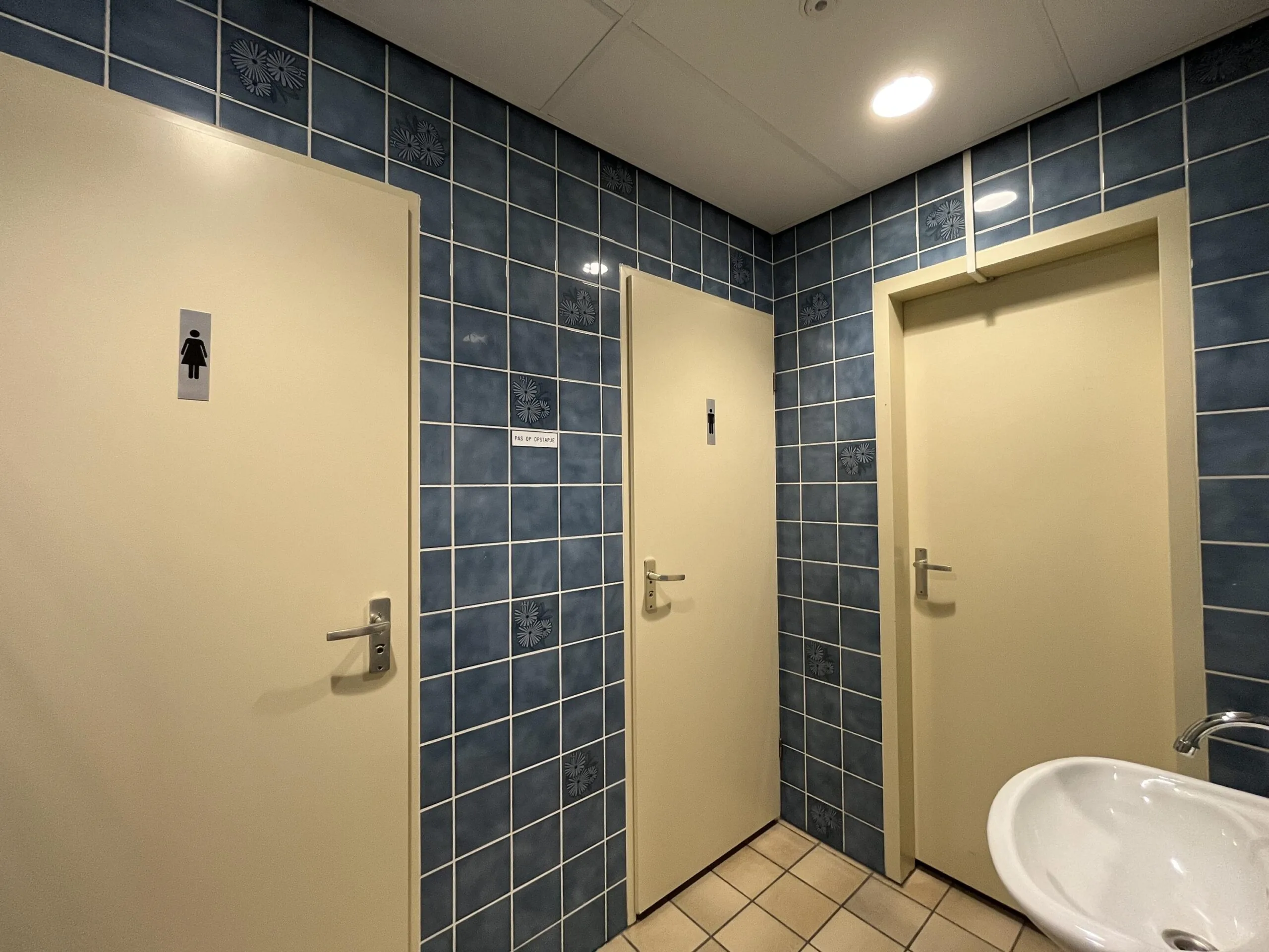 Drie gesloten deuren in een betegelde toiletruimte, waarvan twee met pictogrammen voor mannen en vrouwen.