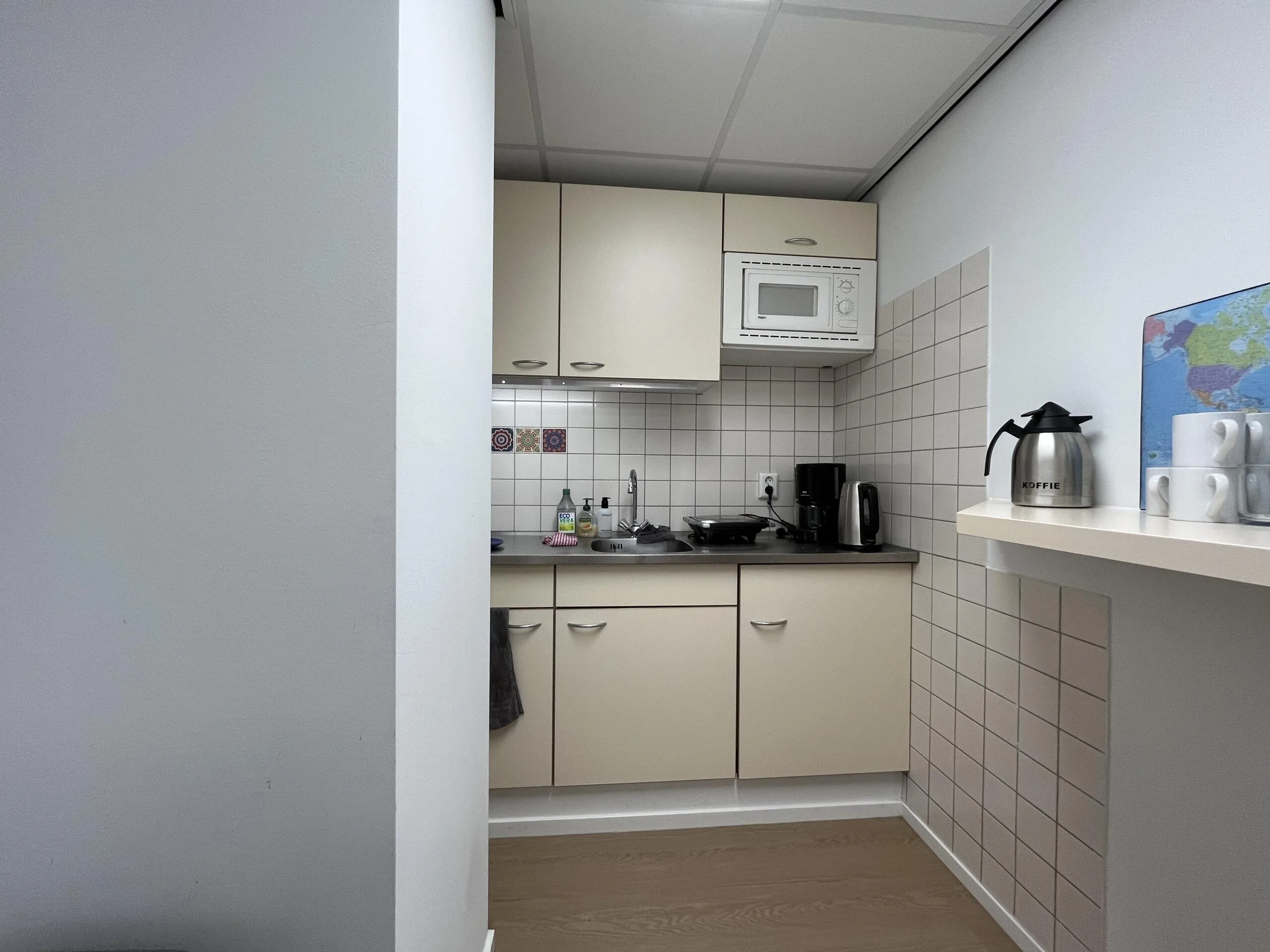 Een kleine keuken in een kantoor of appartement, met beige kasten, een magnetron, koffiezetapparaat en keukengerei.