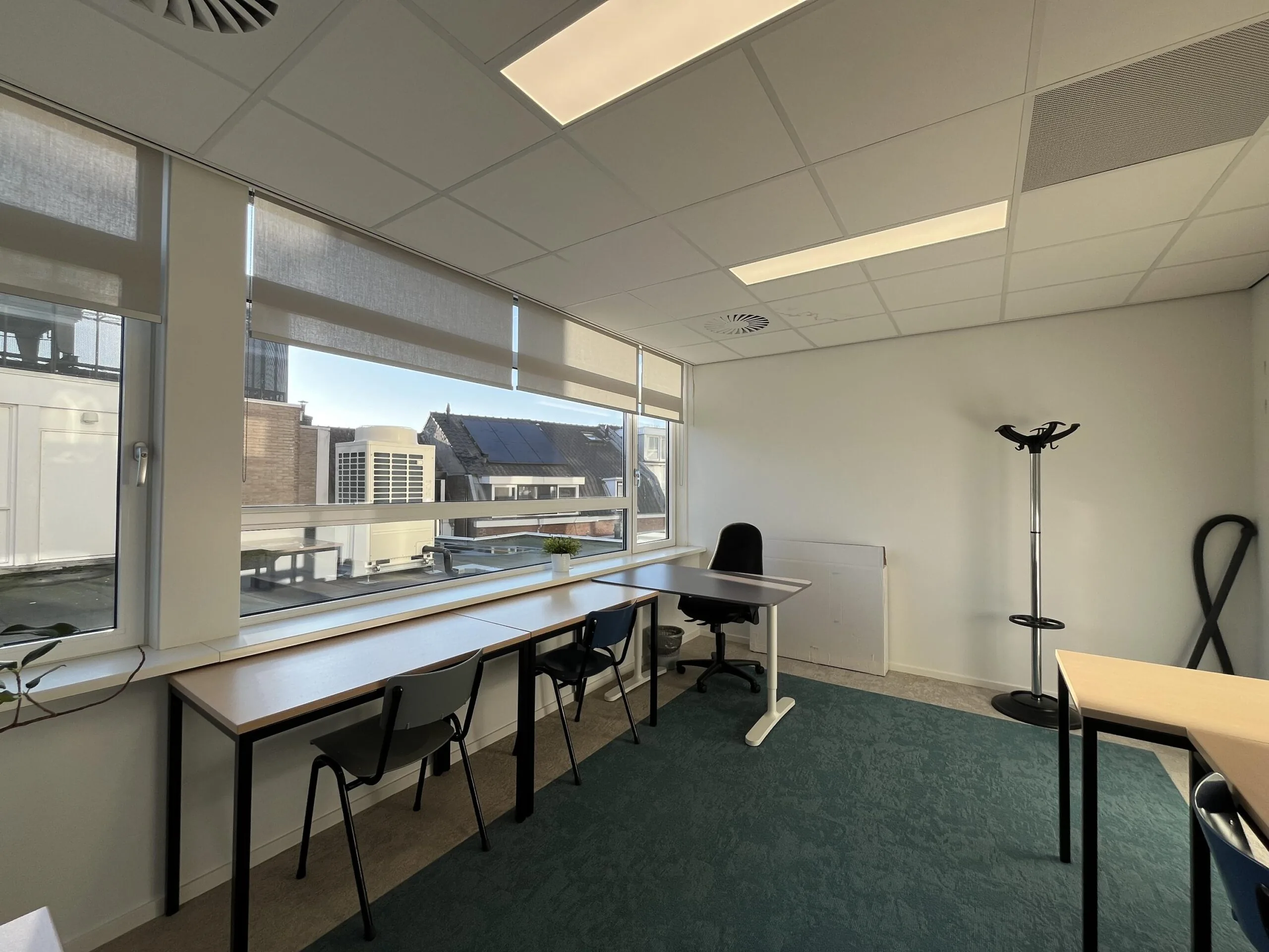 Een moderne kantoorruimte met bureaus, stoelen en een kapstok, met uitzicht op zonnepanelen op nabijgelegen daken.