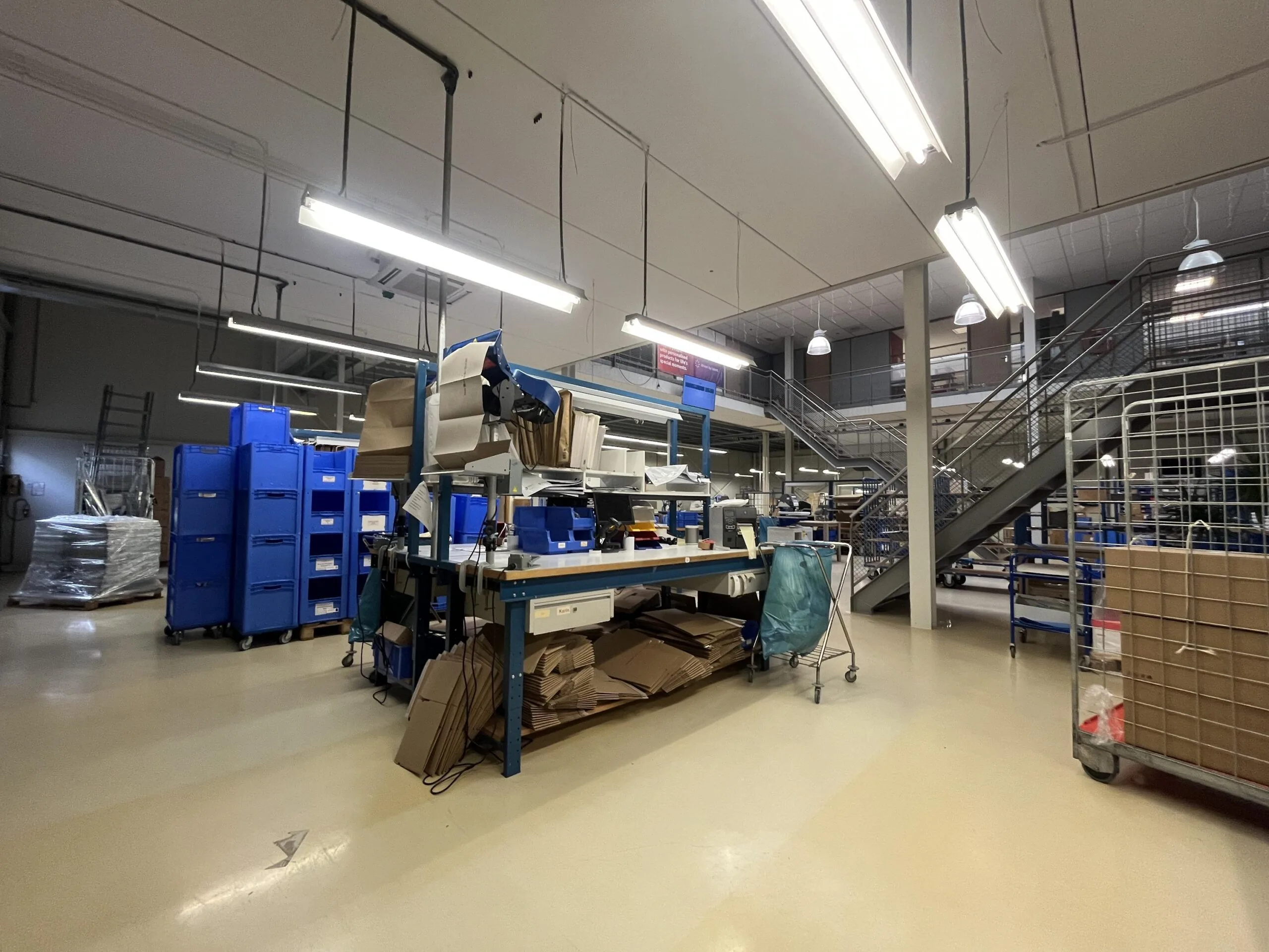 Een magazijn met blauwe kratten, verpakkingsmaterialen en een trap naar een bovenverdieping.
