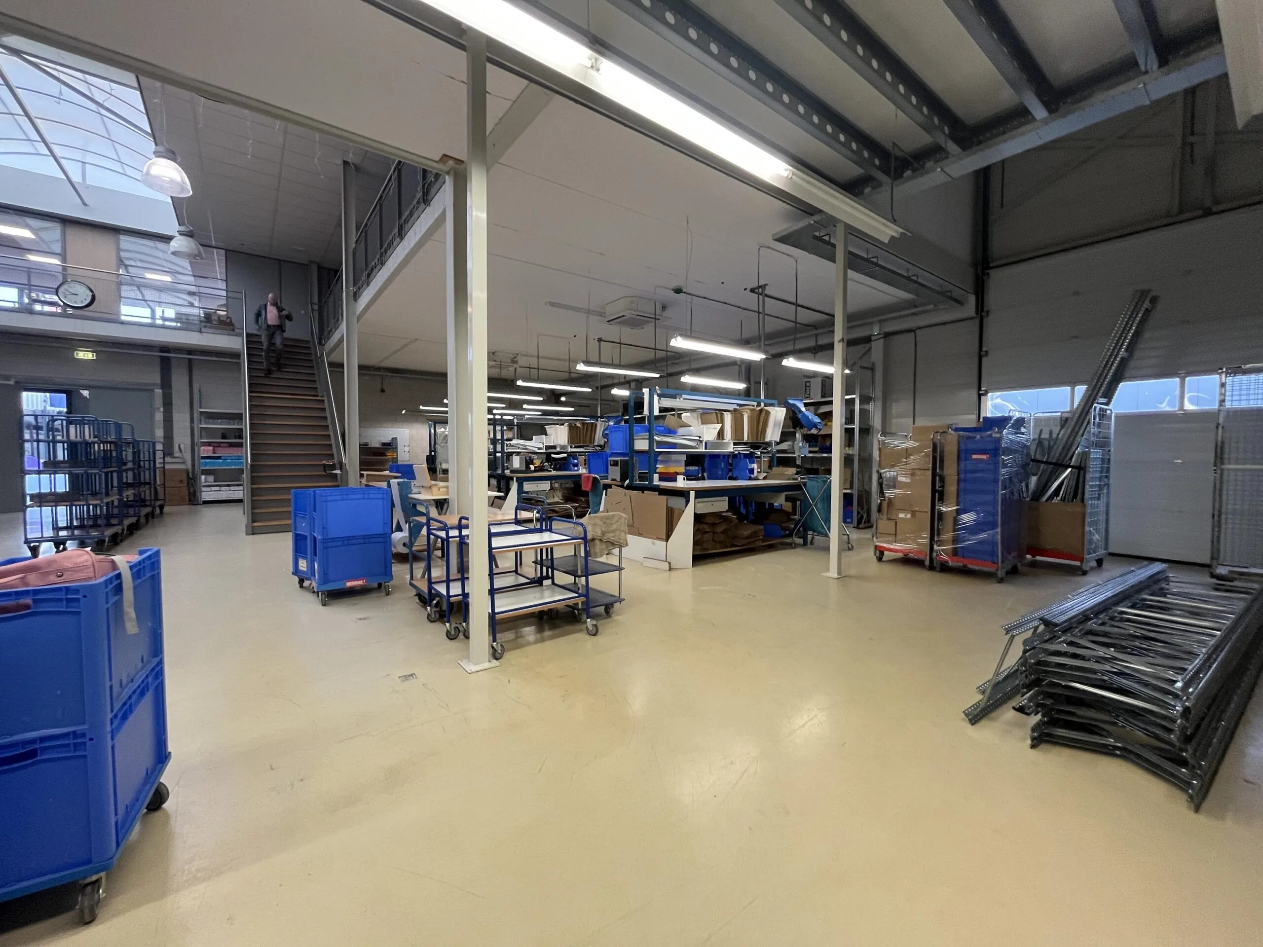 Een magazijn met stellingen, dozen, blauwe rolcontainers en een trap naar een verdieping.