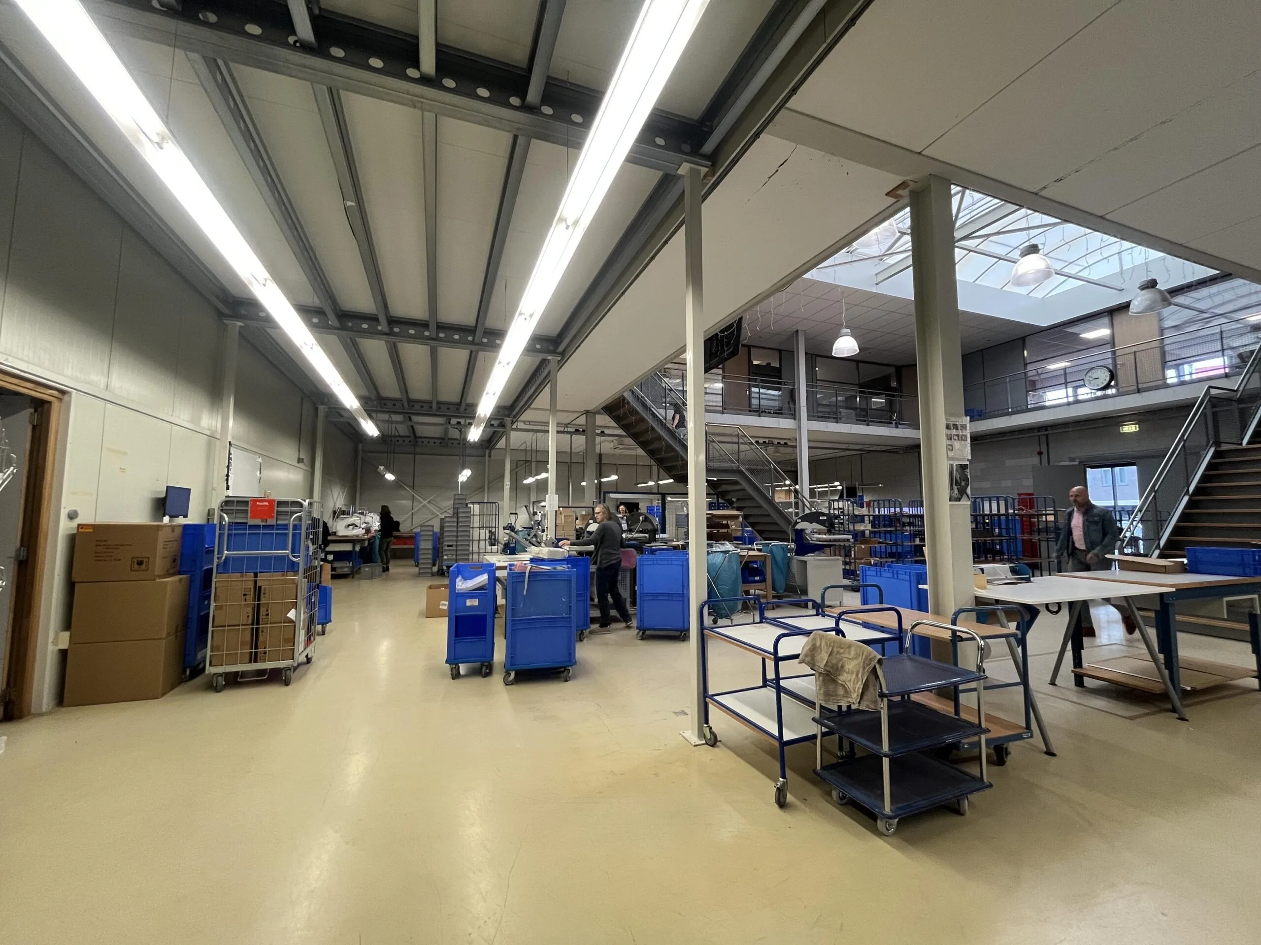 Een magazijn met blauwe karren, dozen en stellingen onder een hoog plafond met tl-verlichting.