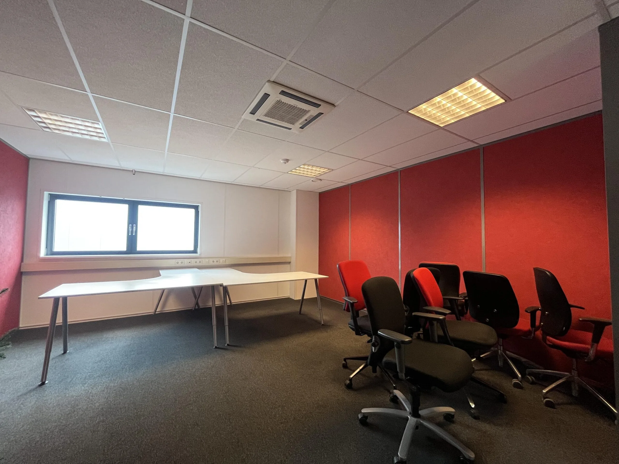 Een vergaderruimte met rode muren, lege witte tafels en meerdere bureaustoelen.