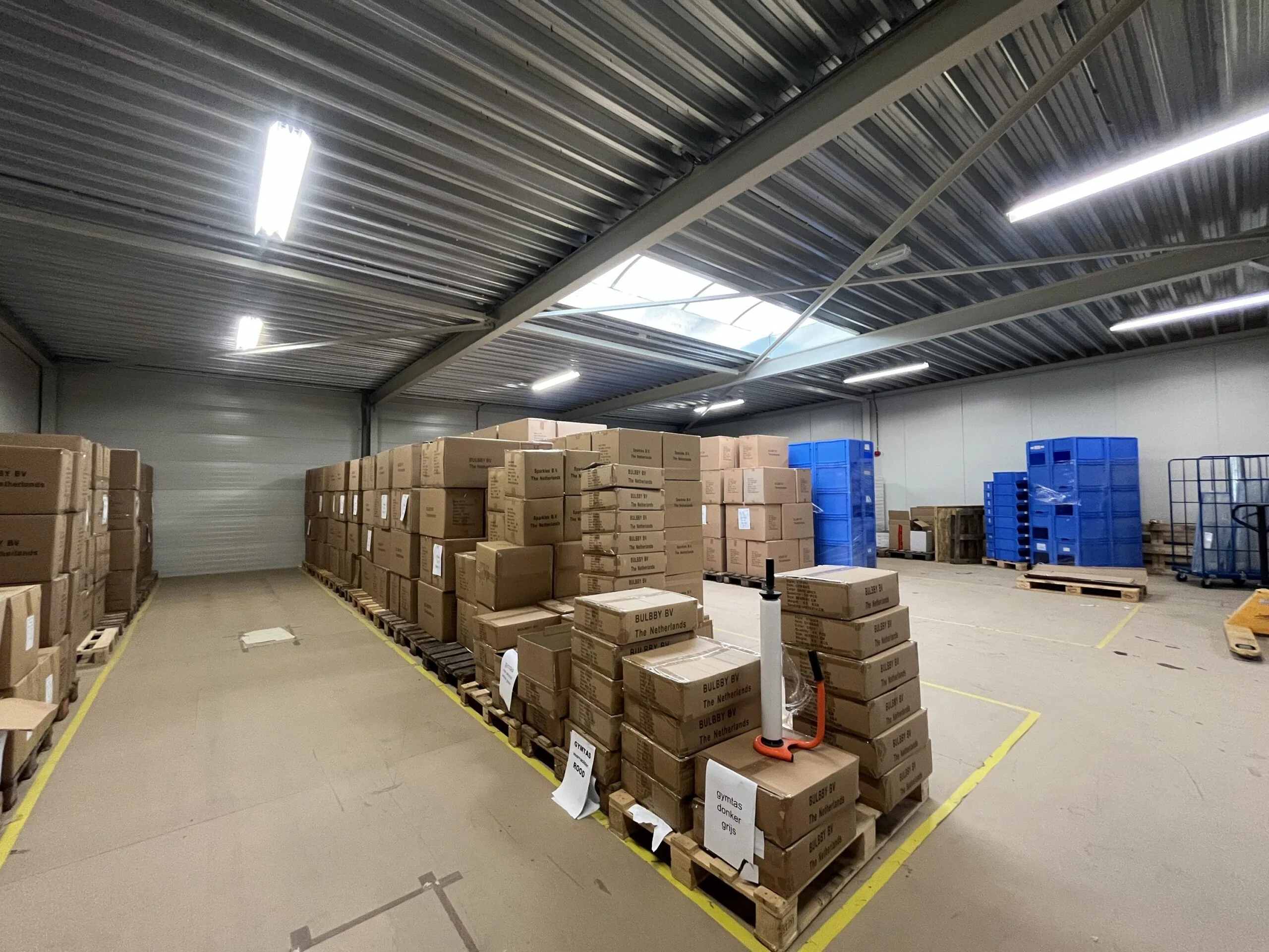 Een opslagruimte met dozen opgestapeld op pallets en blauwe plastic containers tegen de muur.
