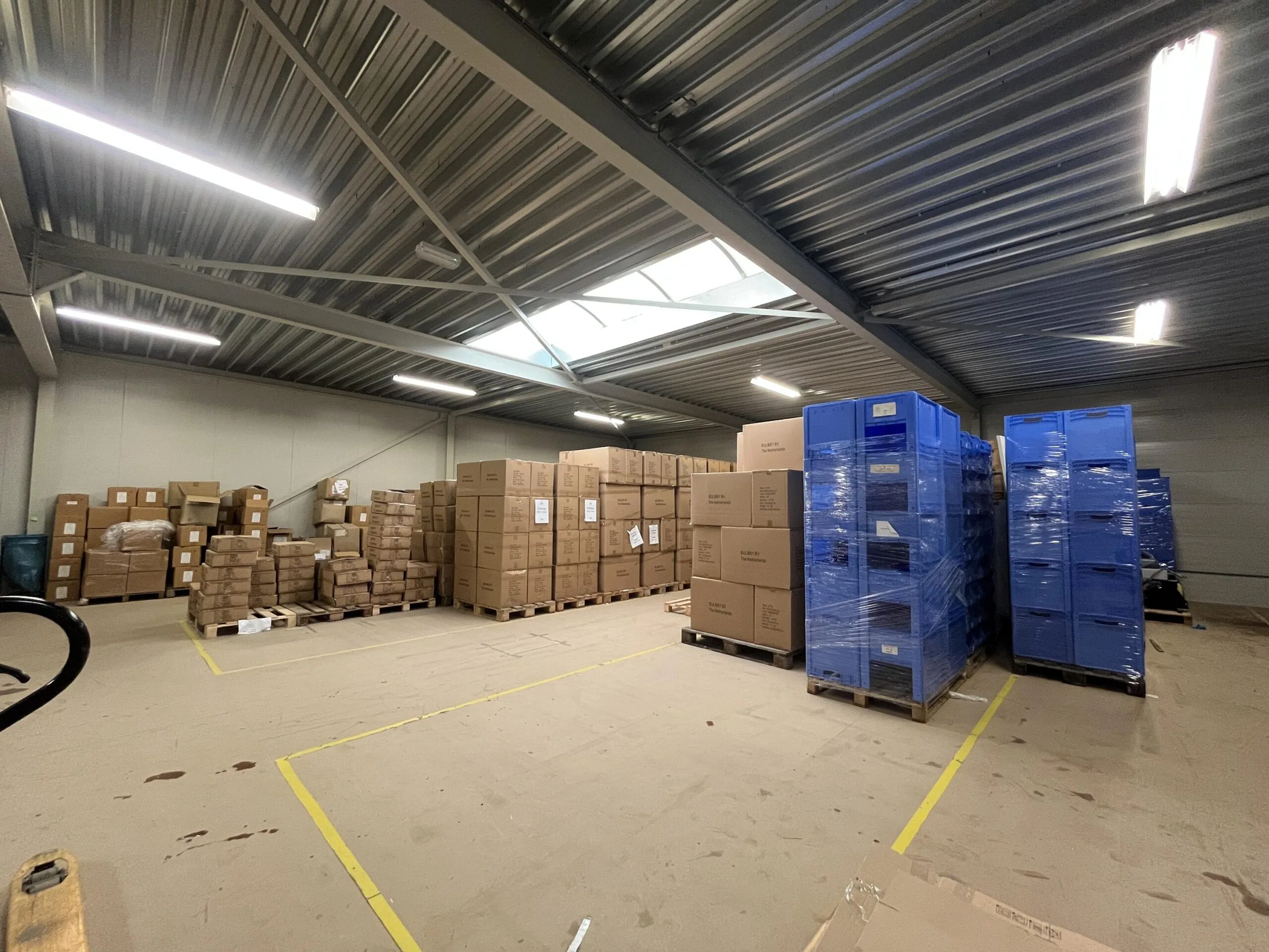 Opslagruimte met gestapelde kartonnen dozen en blauwe plastic kratten op pallets.