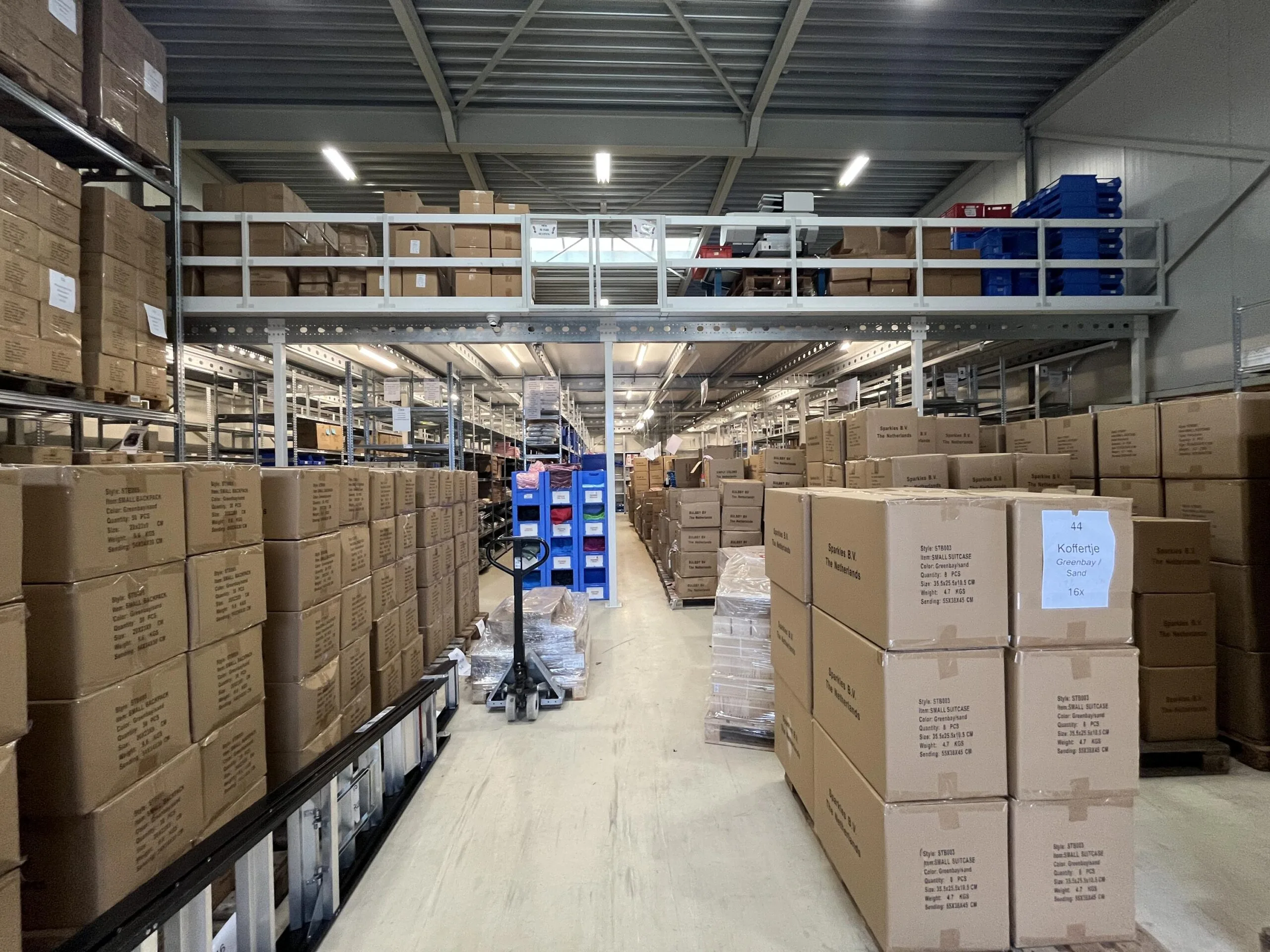 Een magazijn met hoge stellingen vol kartonnen dozen en een palletwagen in het gangpad.