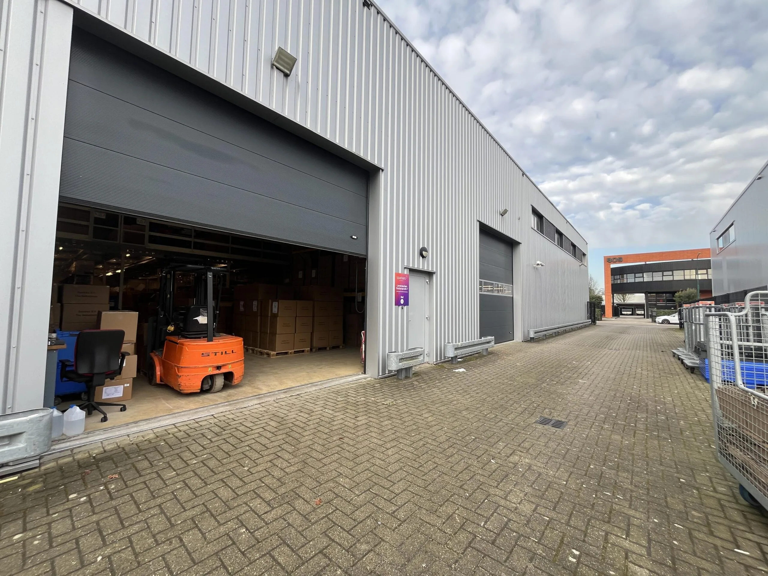 Een buitenaanzicht van een industriële loods met een open roldeur en een oranje heftruck binnenin.