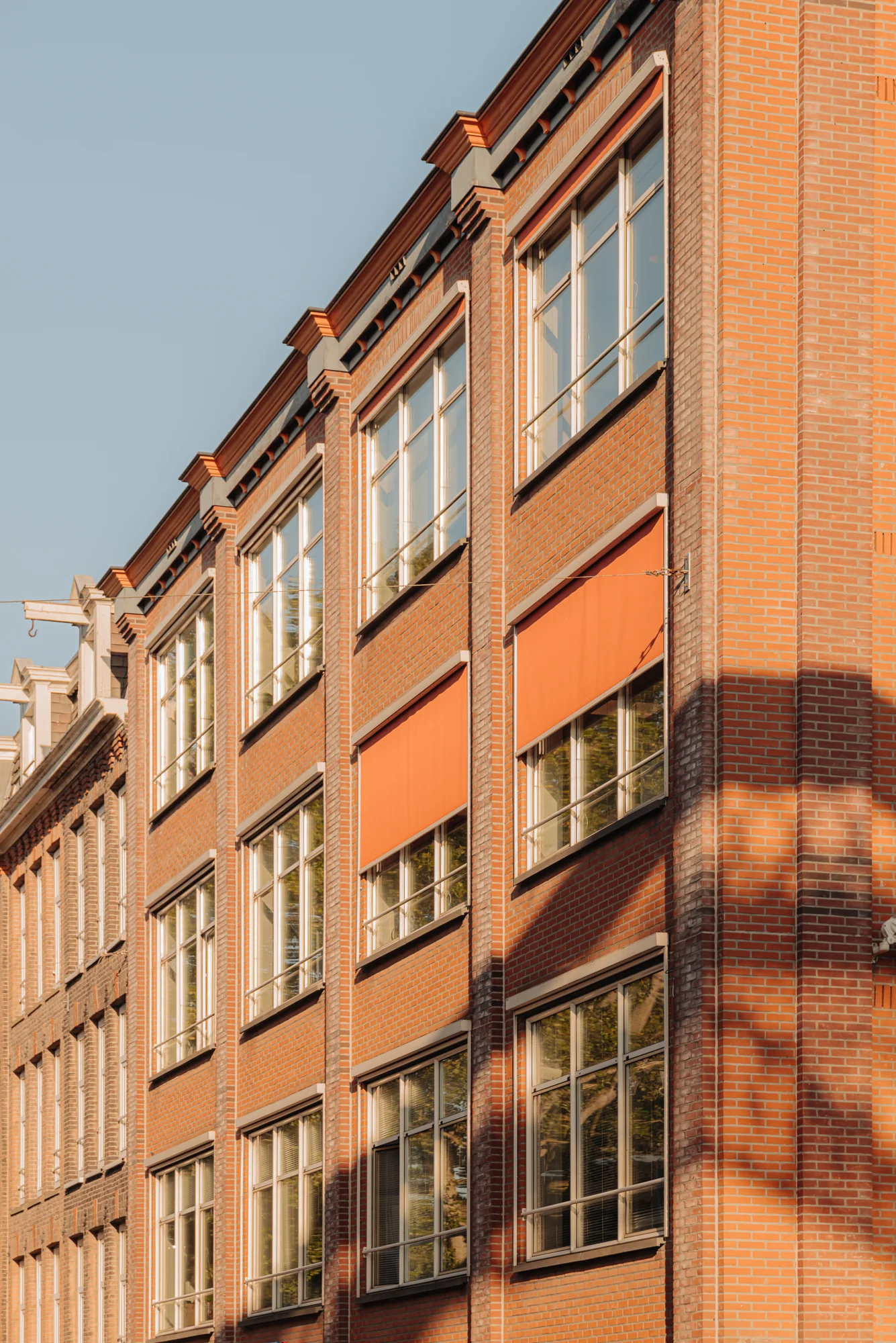 Rood bakstenen gebouw met grote ramen en oranje zonneschermen aan de Hoogte Kadijk.