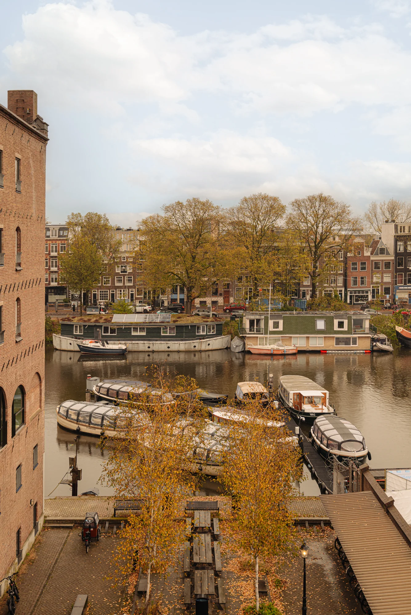 Uitzicht op een gracht met aanlegde boten en woonboten aan de Hoogte Kadijk, omgeven door herfstbomen en oude gebouwen.