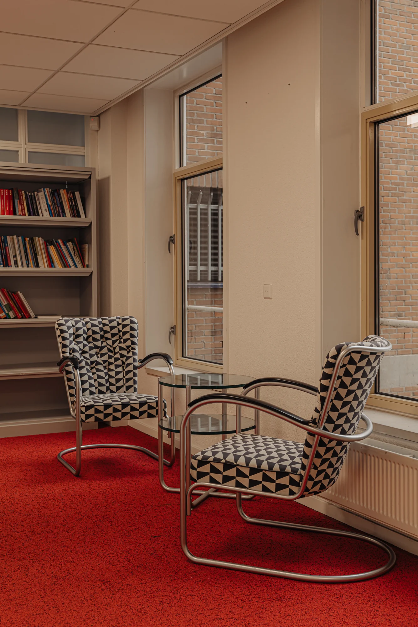 Twee stoelen met geometrisch patroon staan bij een glazen tafel in een kamer met rode vloerbedekking.