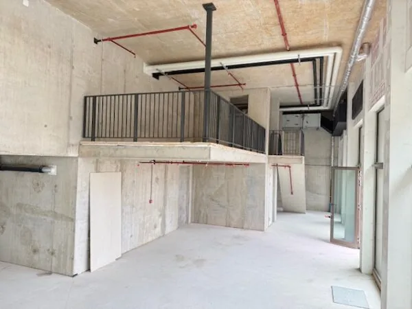 Interieur van een onafgewerkte ruimte met betonnen wanden, een verhoogd platform met zwarte railing, en zichtbare leidingen.